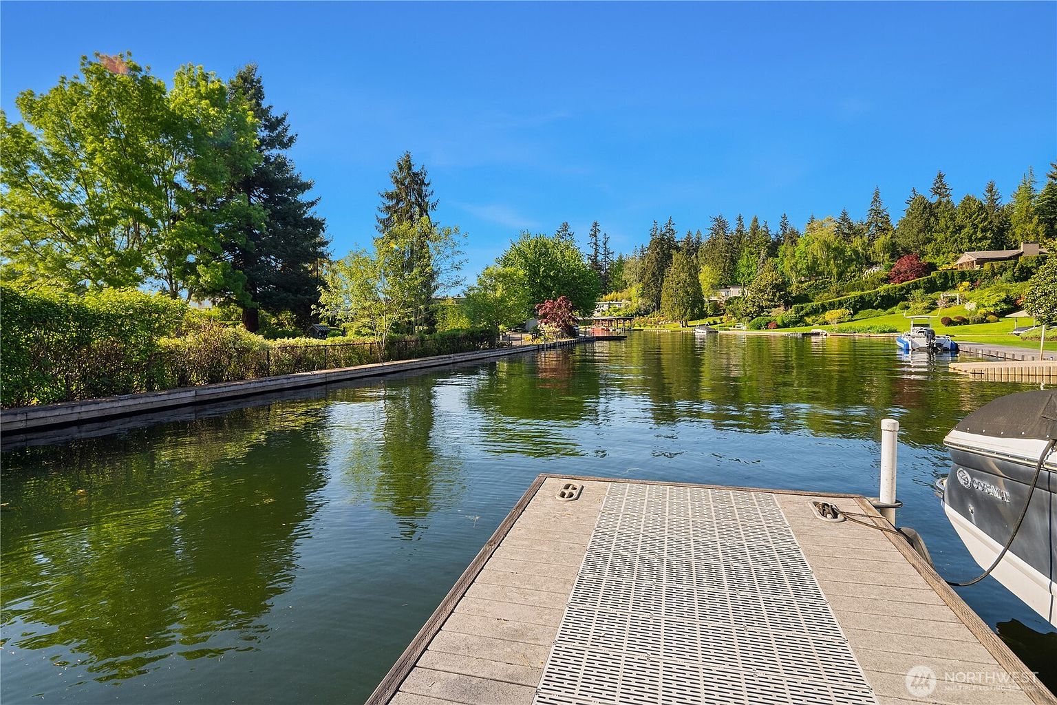 This view captures a serene waterfront setting from the perspective of a private dock extending into a calm canal. The scene features lush green landscaping, mature trees, and a clear blue sky, creating a peaceful and inviting atmosphere. A portion of a boat is visible on the right, emphasizing the property's direct access to the water.