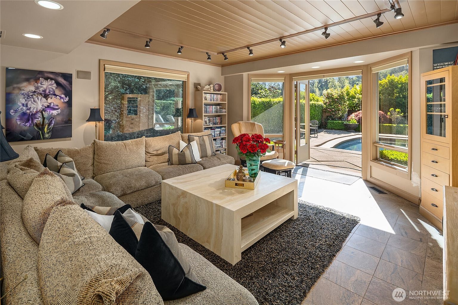 This inviting living room features a large, plush sectional sofa arranged around a substantial light-toned stone coffee table. The space is highlighted by a wood-paneled ceiling with track lighting and large windows that provide a seamless transition to the outdoor pool area. A dark area rug anchors the seating area, while a tall bookshelf and a vibrant floral painting add character to the room.