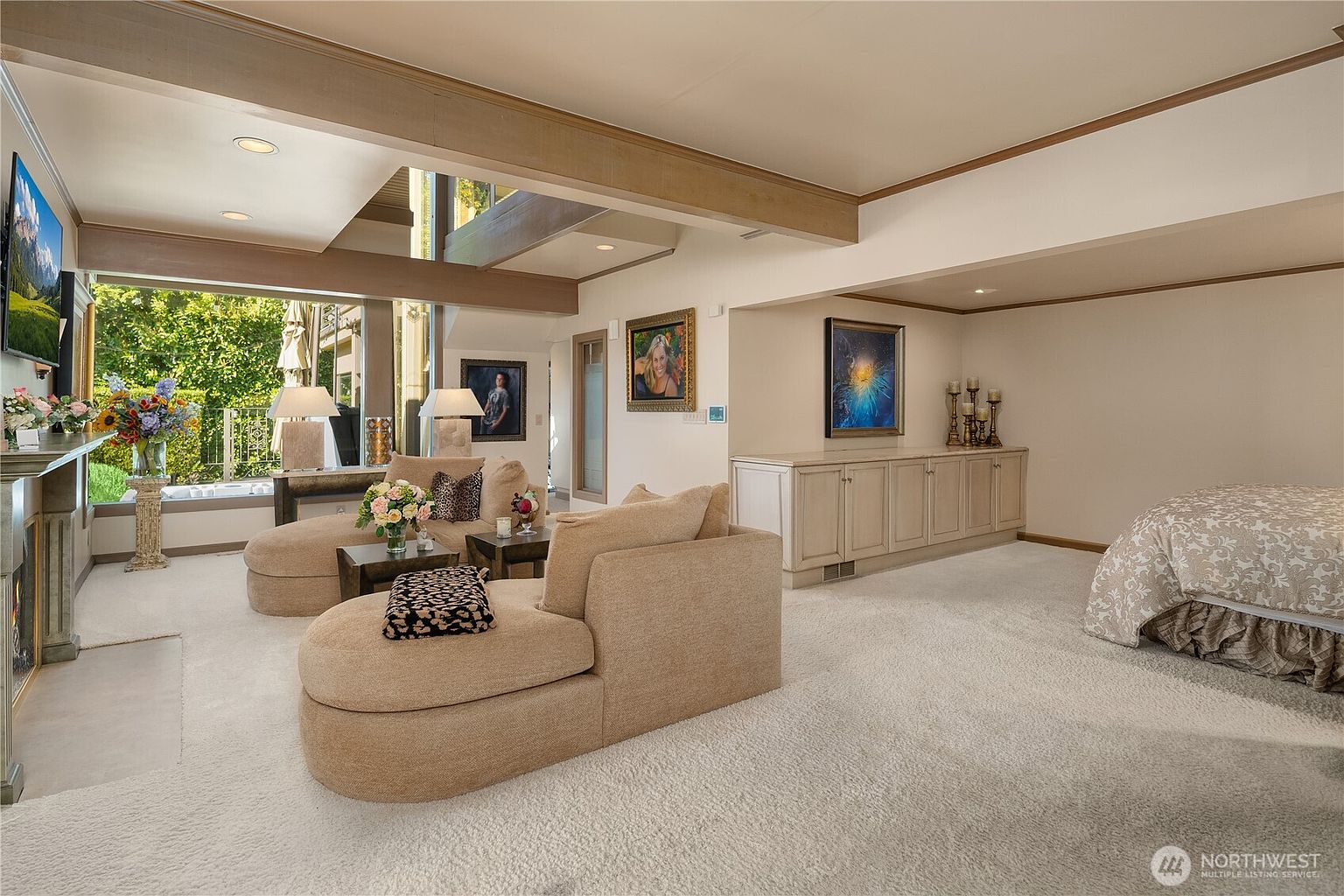 This spacious living area features a neutral color palette with plush, light-toned seating arranged around a central coffee table. A large window wall provides an expansive view of the lush greenery outside, while a built-in cabinet unit offers ample storage along the side wall. The room is accented with framed artwork and decorative lighting, creating a comfortable and inviting atmosphere.