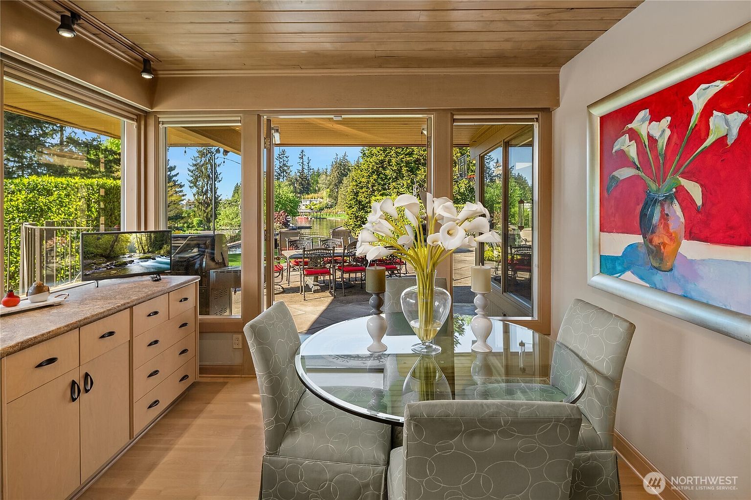 This bright dining area features a glass-topped table surrounded by patterned chairs, positioned in front of large windows that offer a view of an outdoor patio and a lake. To the left, a light wood cabinet provides storage and surface space, while a vibrant painting with white flowers adds a bold pop of color to the wall. The room is finished with a warm wood-paneled ceiling, creating a cozy and inviting atmosphere that seamlessly connects indoor and outdoor living.