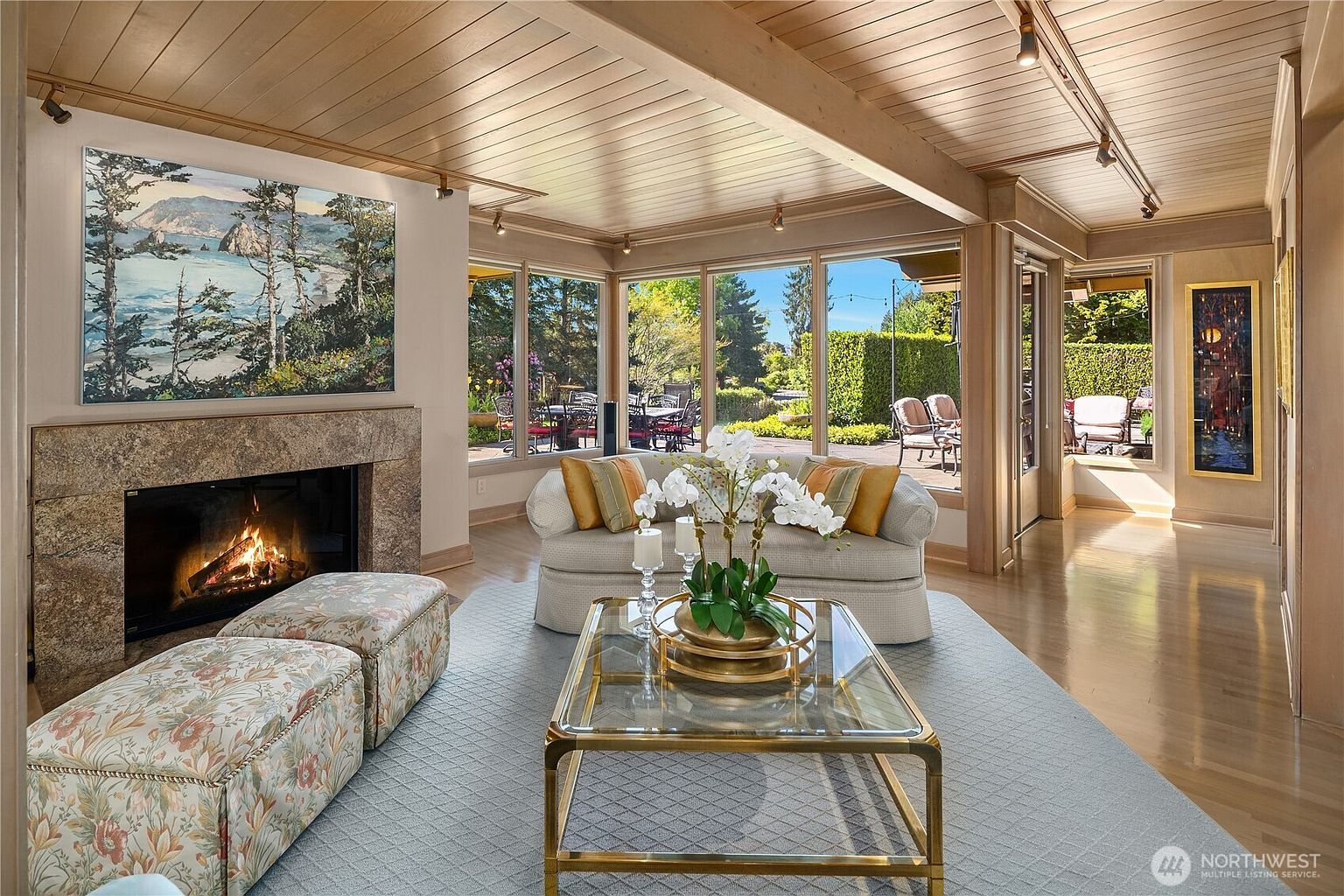 This inviting living room features a warm, wood-paneled ceiling with exposed beams and a stone-clad fireplace surround. A plush sofa and two patterned ottomans are arranged around a glass-topped coffee table, while large windows provide an expansive view of the outdoor patio and lush greenery. The space is decorated with a large landscape painting and elegant floral arrangements, creating a sophisticated and comfortable atmosphere.
