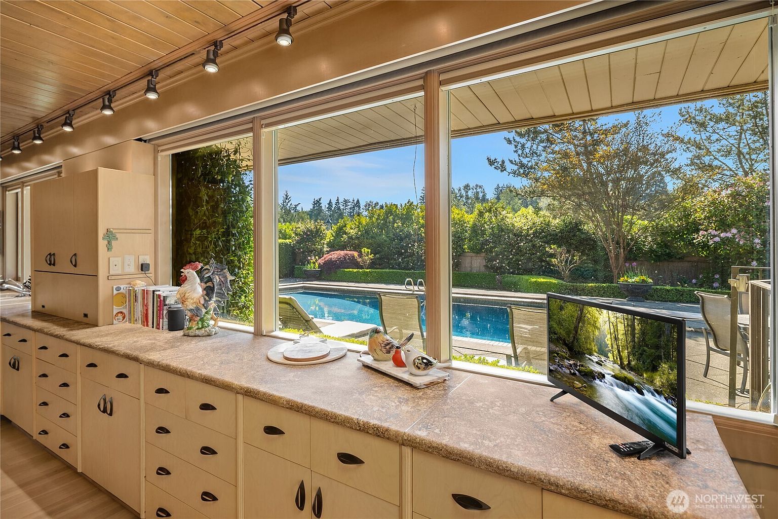 This interior view captures a kitchen counter area featuring light wood cabinetry and a textured stone-look countertop. A long row of large windows provides a clear, expansive view of a backyard swimming pool and lush landscaping, creating a bright and airy atmosphere. The scene is accented by decorative figurines on the counter and a small television, emphasizing a comfortable, lived-in living space.