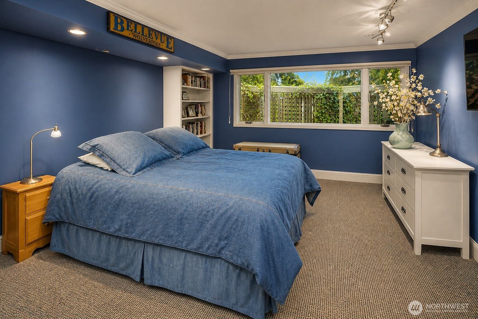 This bedroom features deep blue walls and a large bed with a matching denim-textured comforter. A white bookshelf is built into the wall, and a white dresser with a lamp and floral arrangement sits beneath a wide window that looks out onto a green, leafy backyard. The room is carpeted and illuminated by recessed lighting and a bedside lamp, creating a cozy and personalized atmosphere.
