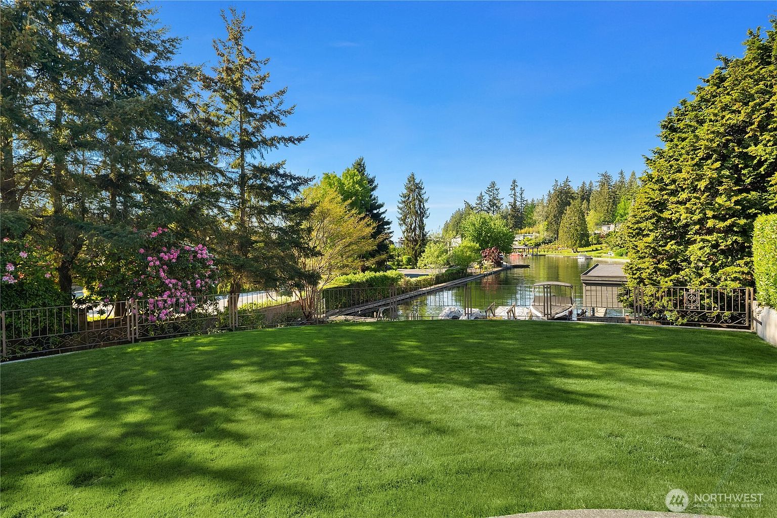 This expansive, manicured lawn serves as a serene backyard overlooking a private waterfront canal. The scene is framed by lush green trees and a vibrant flowering bush, with a metal fence separating the yard from the water where a boat dock is visible. The perspective is a wide, eye-level shot capturing the peaceful transition from the private garden to the tranquil waterway under a clear blue sky.