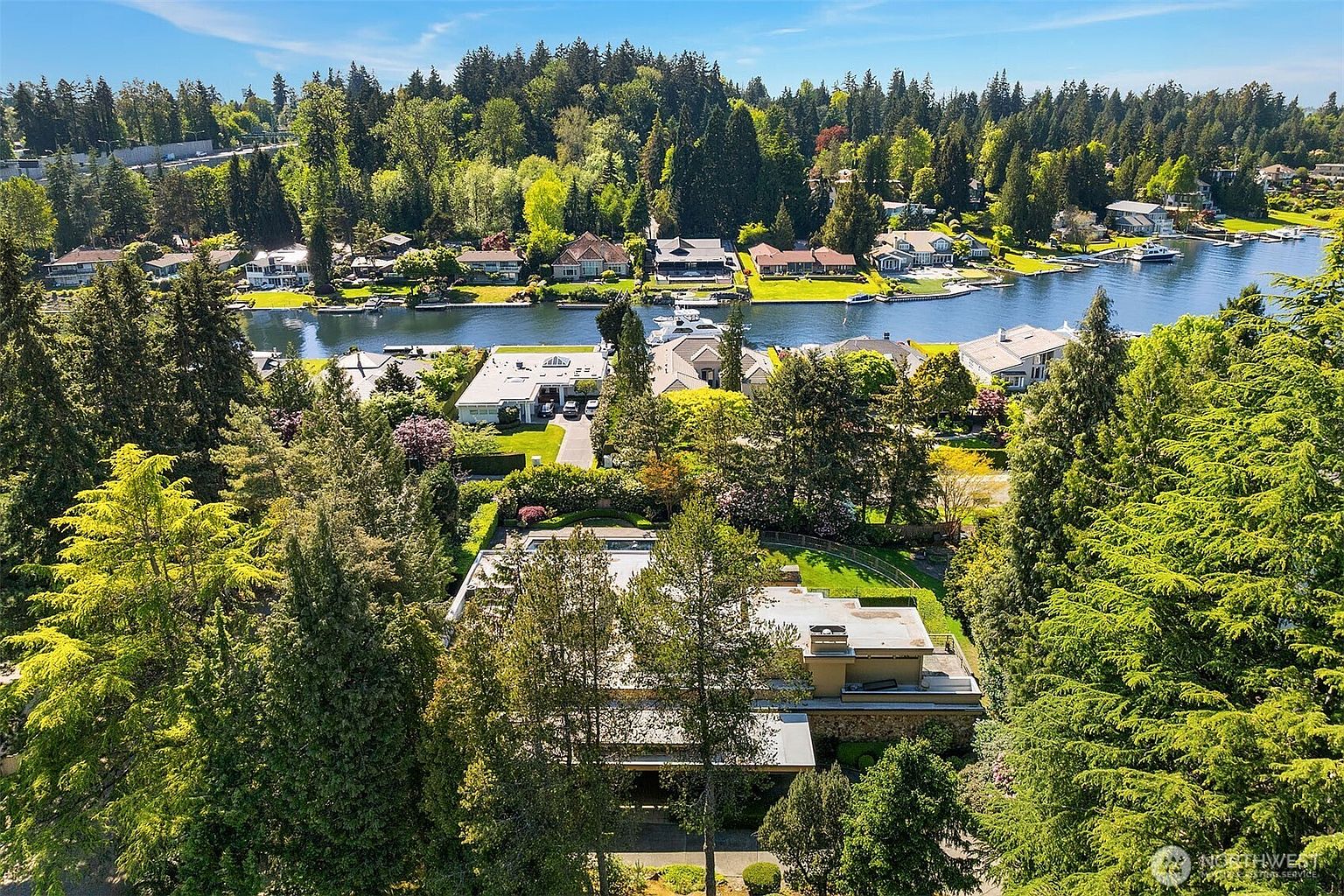 This high-angle aerial perspective captures a sprawling residential property nestled within a lush, tree-filled landscape. The home features a flat-roofed architectural design with stone accents, surrounded by mature greenery and a private driveway, while the background showcases a serene waterway lined with neighboring waterfront homes. The composition emphasizes the property's secluded, park-like setting and its proximity to the scenic canal.