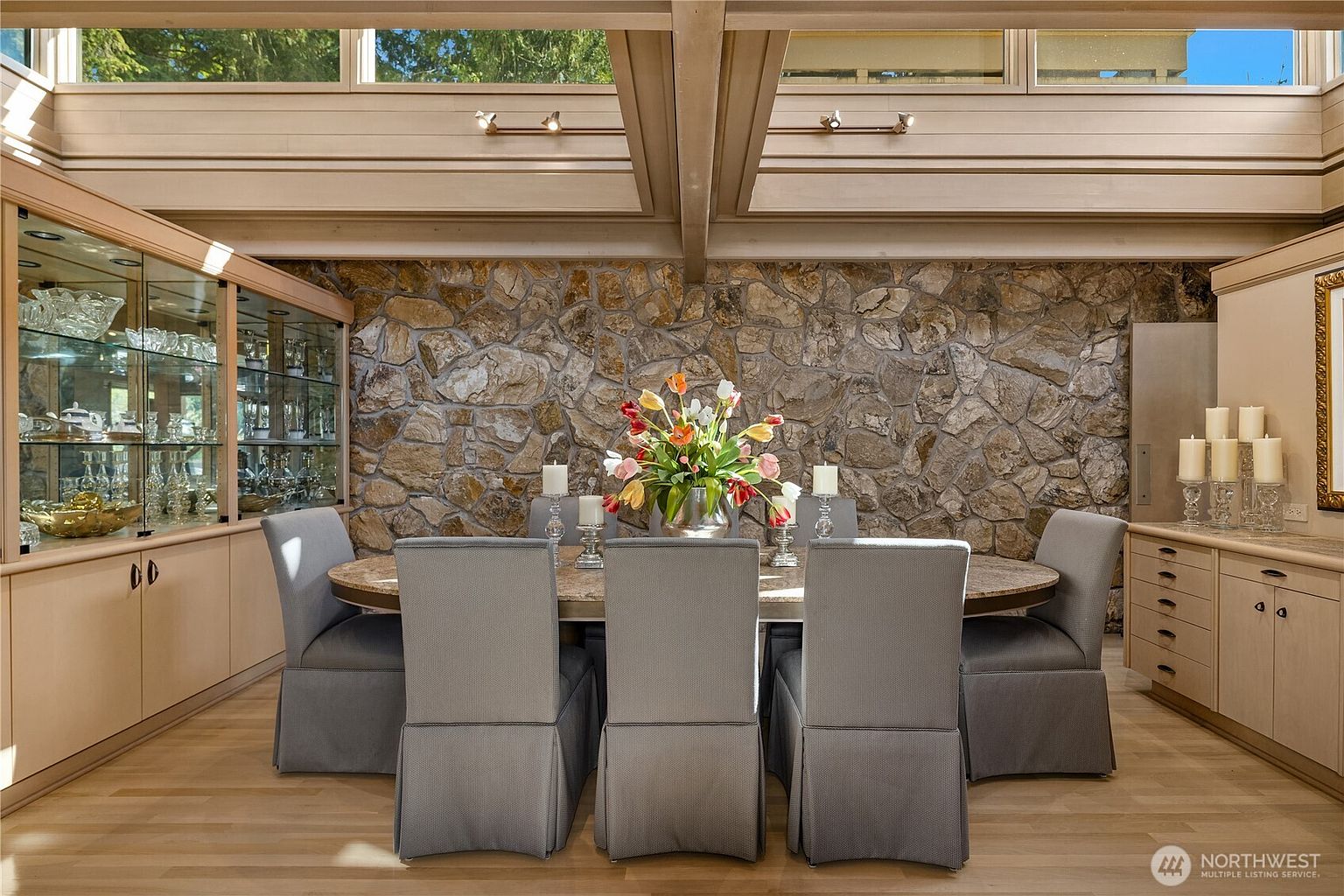 This elegant dining room features a central oval table surrounded by six upholstered chairs, set against a prominent stone-clad wall. The space is illuminated by natural light from a series of clerestory windows above, complemented by built-in glass display cabinets and light wood cabinetry. The room offers a sophisticated, textured aesthetic with a focus on natural materials and clean, modern lines.