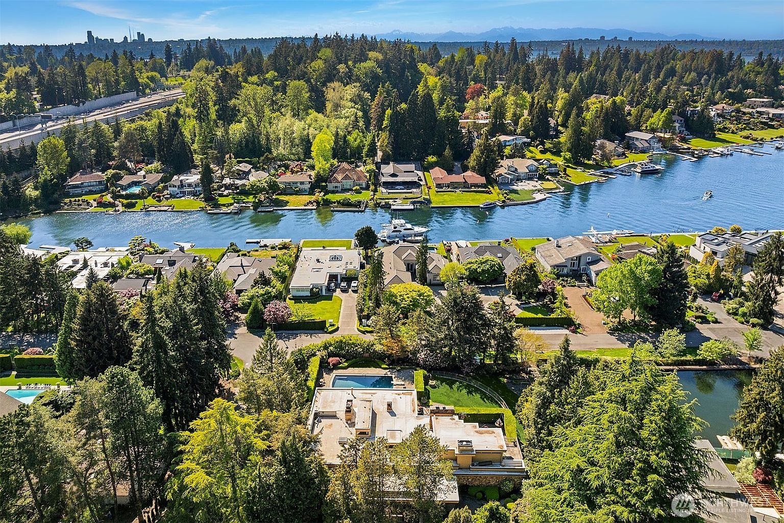 This high-angle aerial view captures a luxurious waterfront residential neighborhood nestled among dense, mature trees. The scene features multiple multi-story homes with private docks, swimming pools, and manicured landscaping along a calm waterway, with a distant city skyline visible on the horizon. The perspective provides a comprehensive look at the property's prime location and its integration into the lush, natural surroundings.