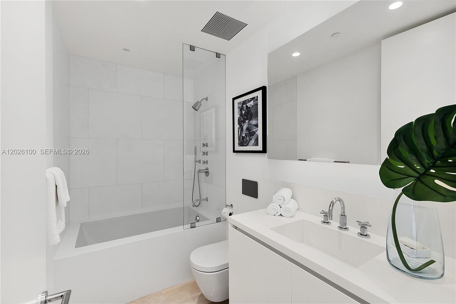 This modern, minimalist bathroom features a sleek white vanity with a rectangular sink and polished chrome fixtures. A glass-enclosed shower and tub combination is tiled in white, complemented by a large, frameless mirror and a framed black-and-white photograph on the wall. The space is bright and clean, accented by a decorative plant and neatly rolled white towels.