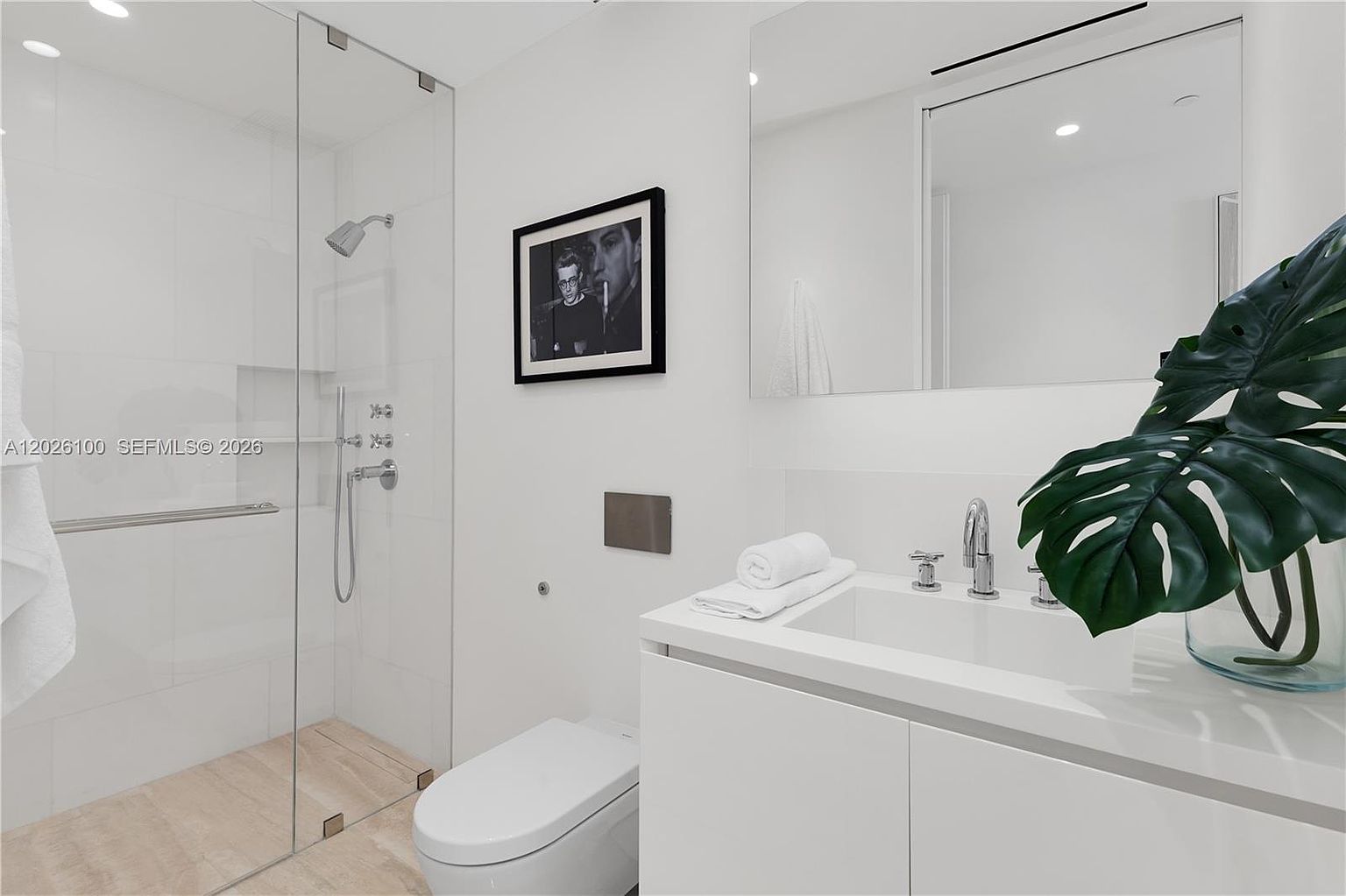 This modern, minimalist bathroom features a sleek white vanity with a rectangular sink and a large, frameless mirror. To the left, a glass-enclosed walk-in shower with light-toned tile flooring adds a clean, airy feel to the space. A framed black-and-white photograph hangs on the wall, providing a sophisticated contrast to the bright, neutral color palette.