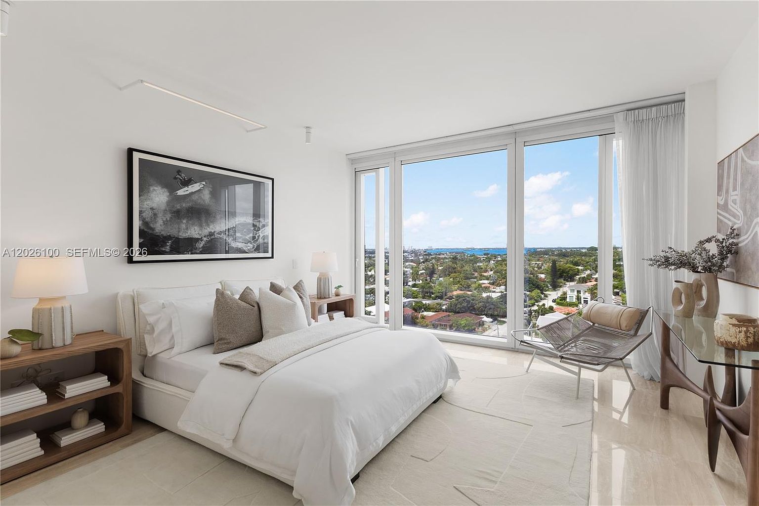 This bright and airy primary bedroom features a neutral color palette and a minimalist aesthetic. The room is anchored by a plush bed with layered pillows and is complemented by a light wood nightstand and a glass-topped console table. Large floor-to-ceiling windows provide an expansive view of the surrounding neighborhood and distant water, filling the space with natural light.