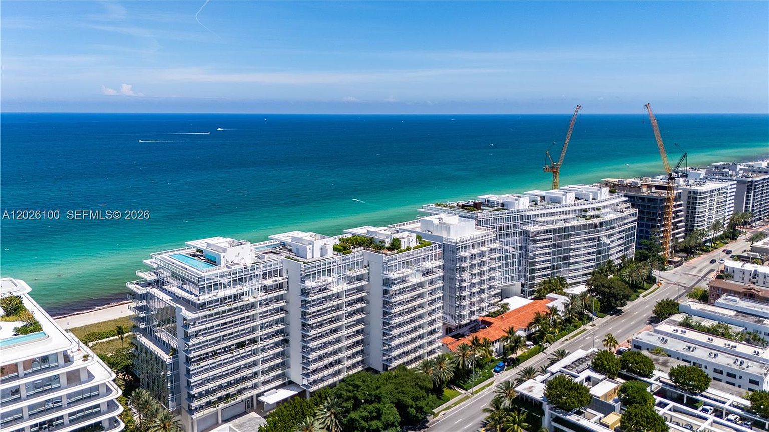 This high-angle aerial view captures a modern, multi-story residential complex situated directly along a pristine coastline. The architecture features clean lines, extensive glass balconies, and rooftop amenities, all set against the backdrop of a vibrant turquoise ocean. The scene includes active construction with cranes visible, highlighting the ongoing development and prime beachfront location of the property.