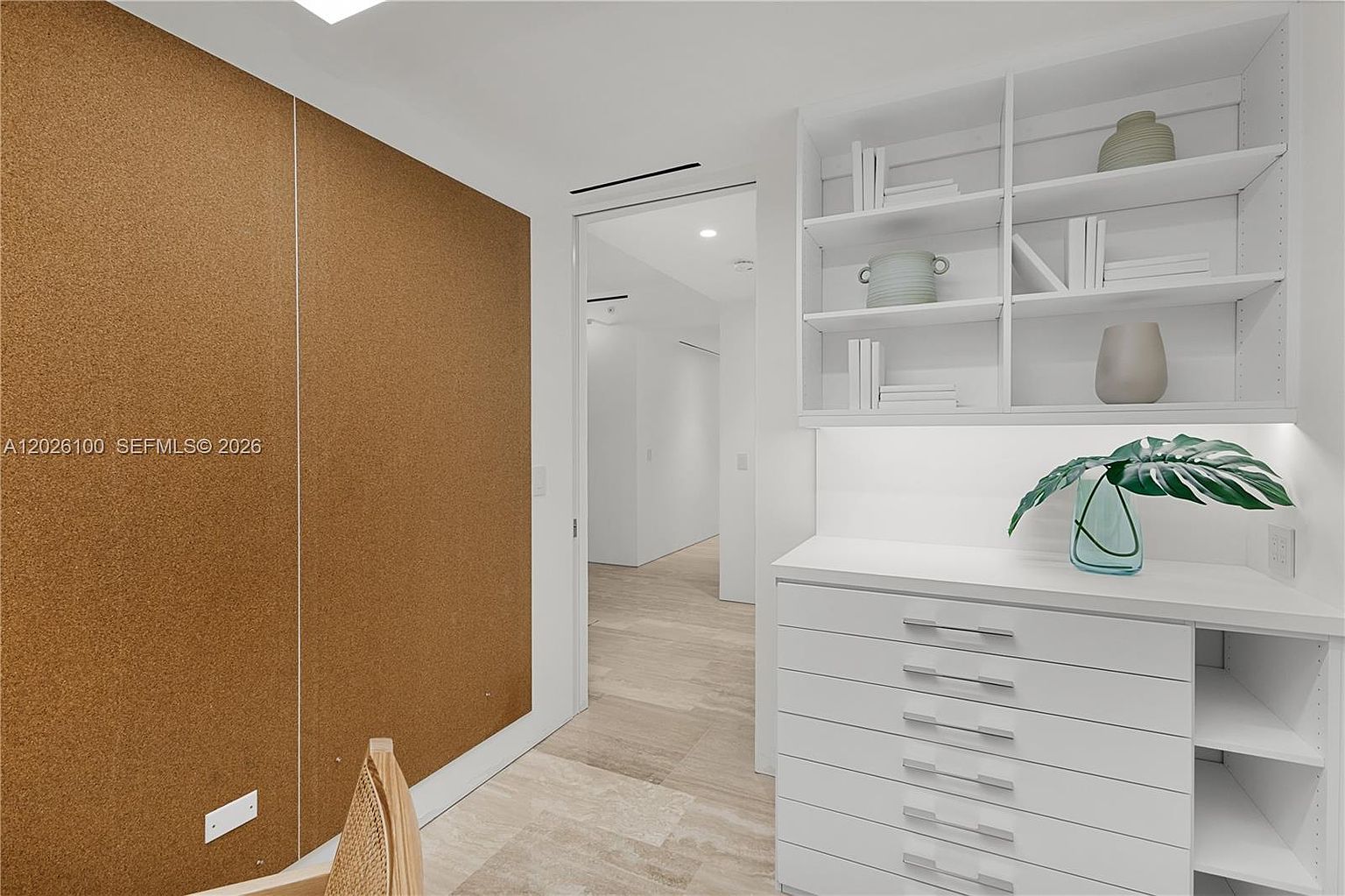 This bright and minimalist home office or study features a large cork-covered accent wall and a built-in white storage unit with open shelving and a bank of drawers. The space is connected to a hallway through a doorway, showcasing light-toned wood flooring throughout. A small potted plant sits on the counter, adding a touch of greenery to the clean, contemporary aesthetic.