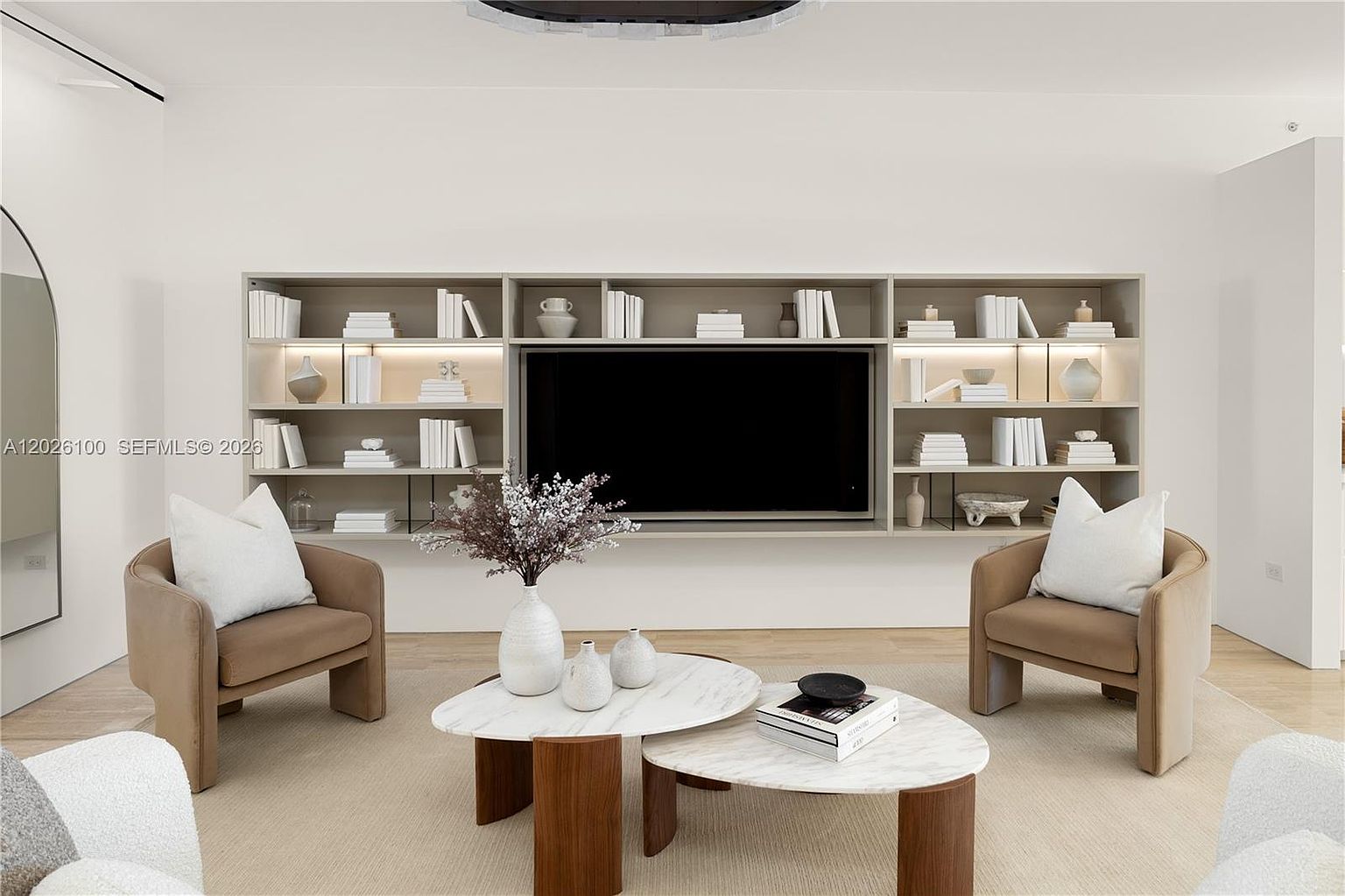 This contemporary living room features a minimalist aesthetic with a large, integrated wall unit housing a television and open shelving for decor. Two plush, neutral-toned armchairs are positioned around a pair of tiered, organic-shaped coffee tables set on a light-colored area rug. The space is bright and airy, characterized by clean lines and a sophisticated, monochromatic color palette.