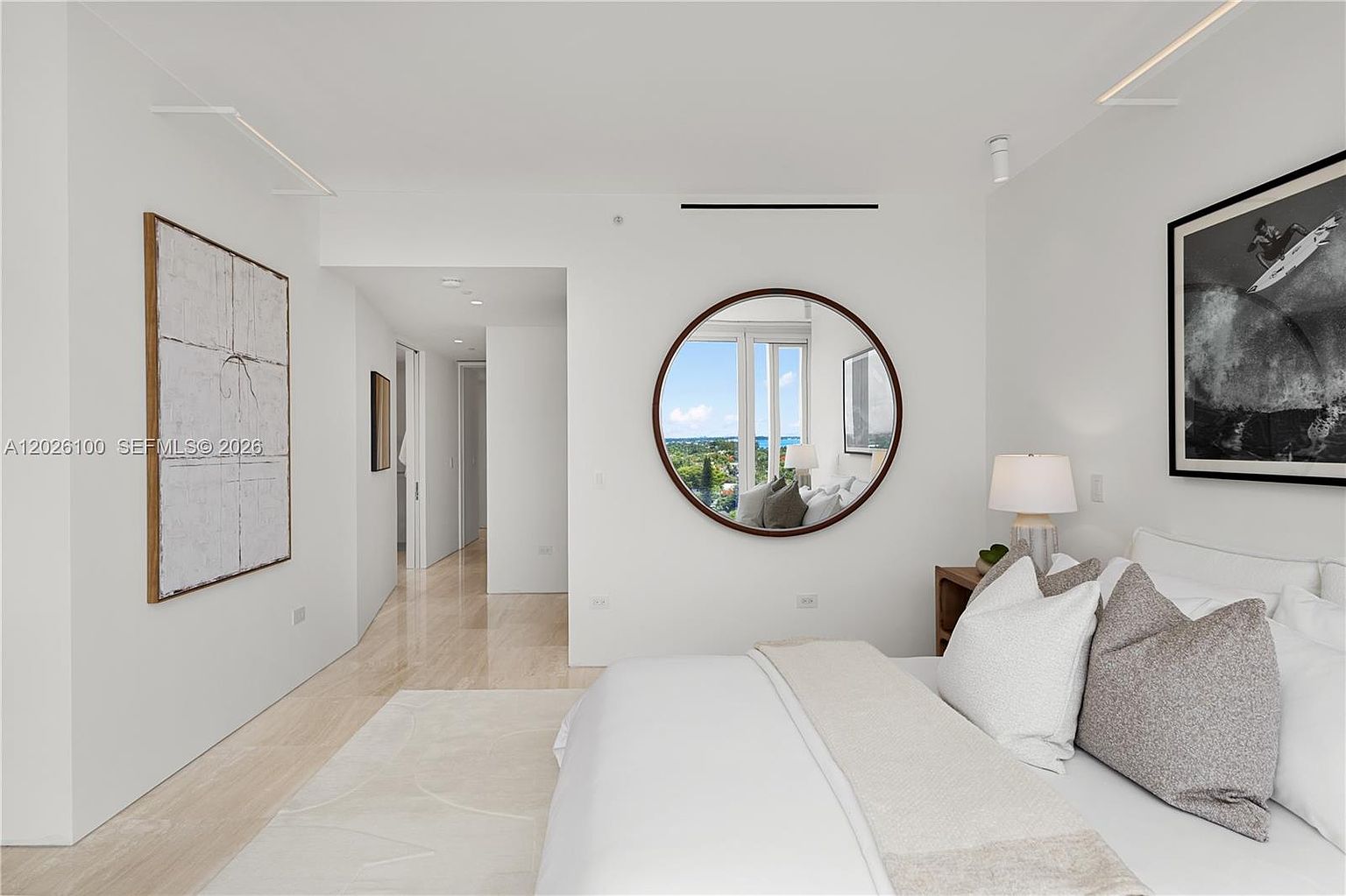 This bright and minimalist primary bedroom features a neutral color palette with white walls and light-toned flooring. A large, round mirror reflects the outdoor view, while a framed black-and-white surf photograph adds a modern artistic touch above the bed. The space is clean and airy, with a hallway leading to other areas of the home, creating a sense of openness and sophisticated simplicity.