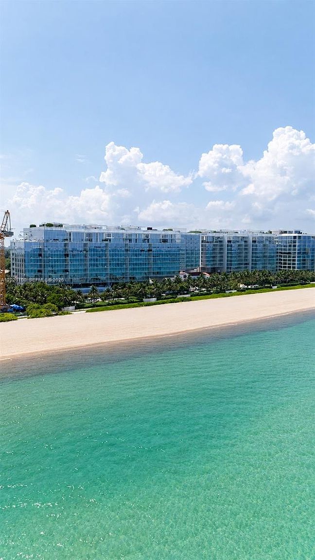 This aerial perspective captures a modern, multi-story residential complex situated directly along a pristine sandy beach. The building features a contemporary design with extensive glass facades that reflect the bright blue sky and surrounding tropical landscape. The turquoise ocean water meets the shoreline in the foreground, creating a serene and luxurious coastal atmosphere.