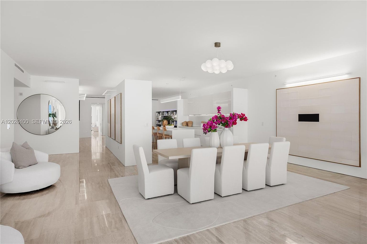 This bright and airy open-concept dining area features a minimalist aesthetic with a light-toned dining table surrounded by six upholstered white chairs. A large, neutral-toned area rug anchors the space, while a modern, cloud-like pendant light fixture hangs above the table. The room flows seamlessly into a sleek, white-cabinet kitchen in the background, and a large circular mirror adds depth to the adjacent living area.