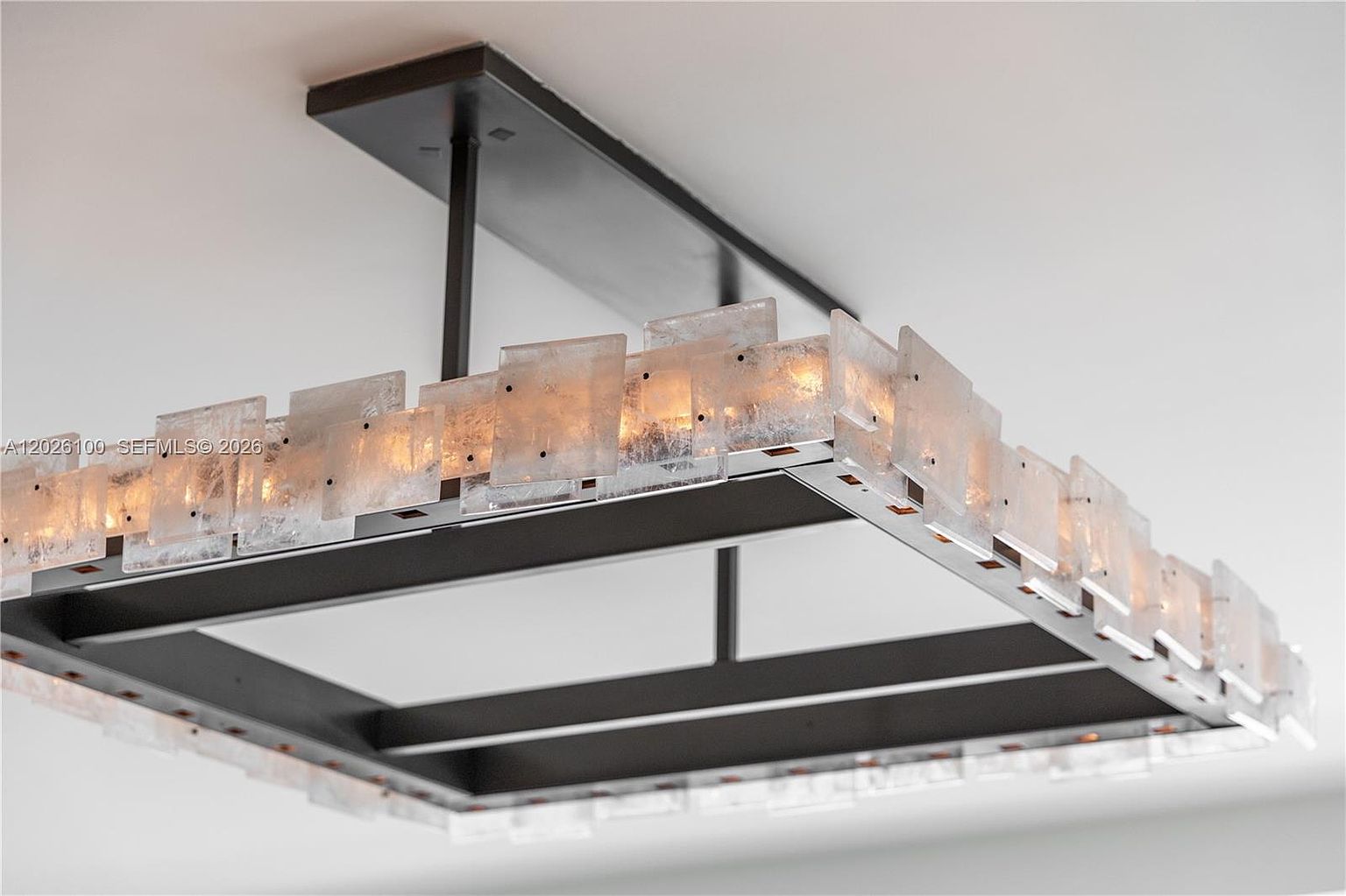 This close-up shot highlights a modern, rectangular chandelier featuring a dark metal frame adorned with numerous textured, translucent glass panels. The fixture is illuminated from within, creating a warm, ambient glow that emphasizes the intricate, frosted quality of the glass. The perspective is angled from below, showcasing the geometric design and the sophisticated, contemporary aesthetic of the lighting piece.