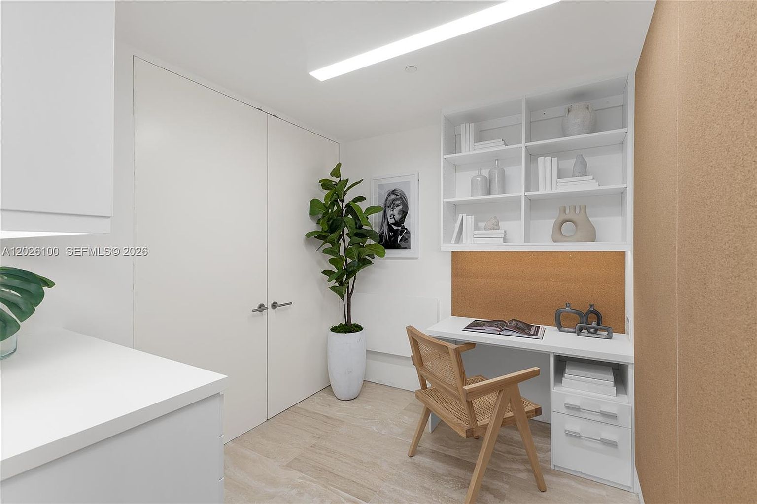 This bright and minimalist home office features a built-in white desk and shelving unit, complemented by a light wood chair and a cork board backsplash. A tall potted plant adds a touch of greenery to the corner, while a framed piece of art hangs on the wall near the double doors. The space is finished with light-toned flooring, creating a clean and productive atmosphere.