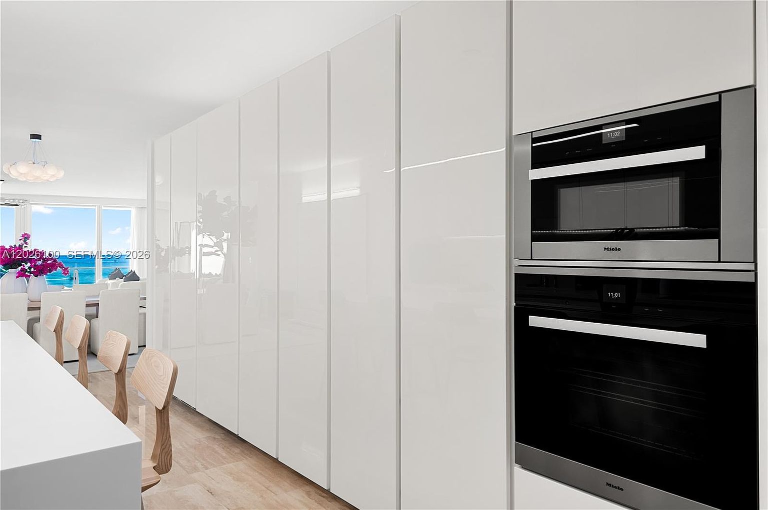 This modern kitchen features a wall of sleek, white, handleless cabinetry that provides a clean and minimalist aesthetic. A built-in Miele oven and microwave stack are integrated into the cabinetry, showcasing high-end appliances. To the left, a dining area with light wood chairs is visible, with large windows in the background offering a bright, airy view of the outdoors.