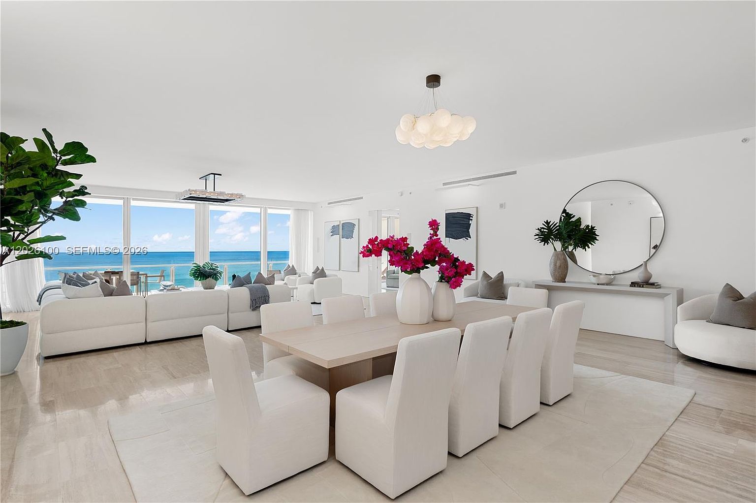 This bright and airy open-concept living and dining area features a sophisticated, minimalist aesthetic with a neutral color palette. A large dining table with eight upholstered chairs serves as the centerpiece, complemented by a modern cloud-like chandelier and vibrant floral arrangements. The space flows seamlessly into a living area with plush white seating, all framed by expansive floor-to-ceiling windows that provide a stunning, unobstructed view of the ocean.