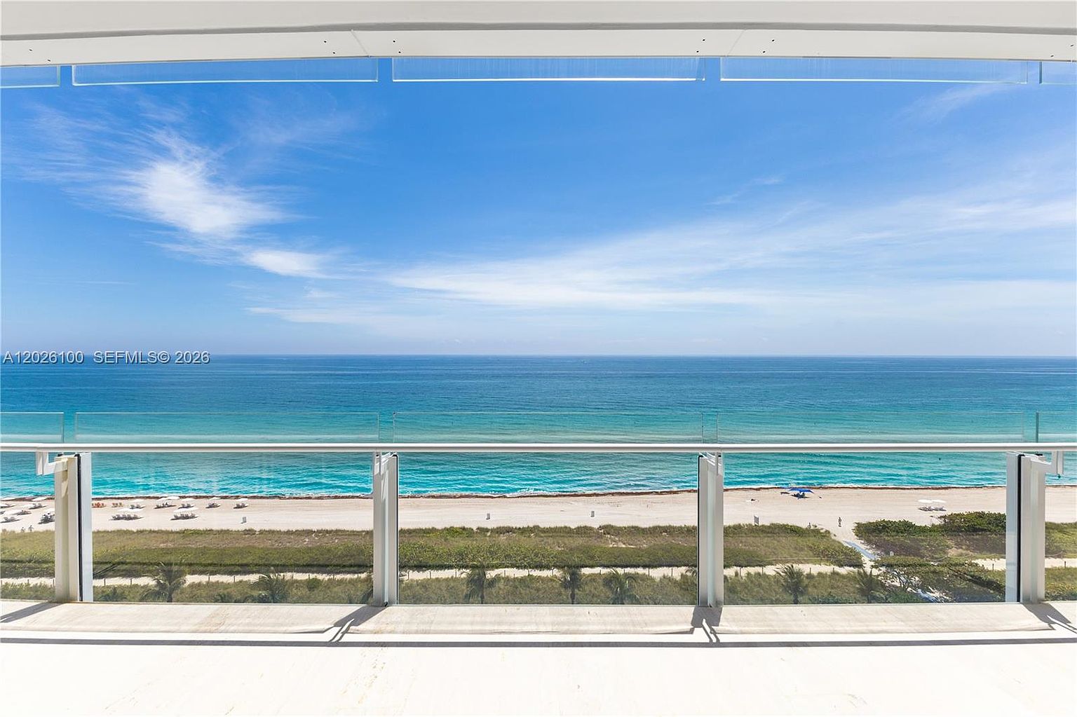 This image captures a direct, eye-level view from a balcony overlooking a pristine beach and the vast ocean. The balcony features a modern glass railing that provides an unobstructed perspective of the turquoise water, sandy shoreline, and coastal vegetation below. The bright, clear sky and expansive horizon create a serene and luxurious atmosphere, emphasizing the property's prime waterfront location.