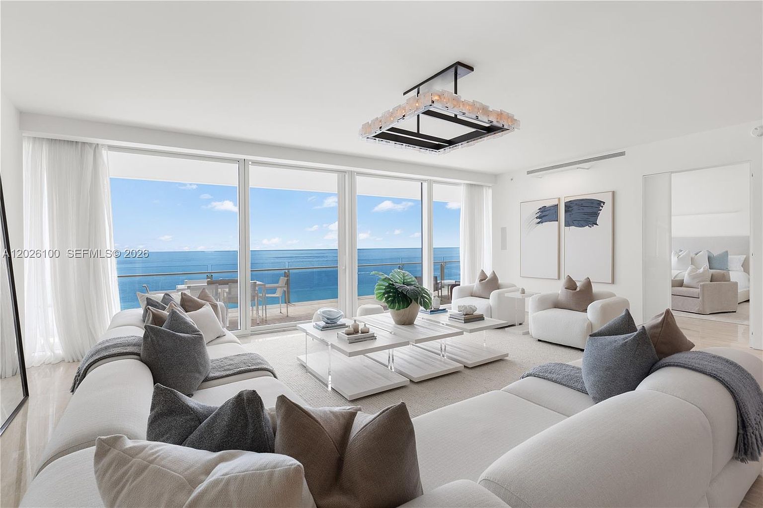 This bright and airy living room features a spacious white sectional sofa and two matching armchairs arranged around a set of tiered, light-toned coffee tables. The room is defined by expansive floor-to-ceiling windows that provide an unobstructed, panoramic view of the ocean and sky. A modern, rectangular chandelier with a geometric frame hangs centrally, while minimalist abstract art adorns the wall, creating a sophisticated and serene coastal atmosphere.