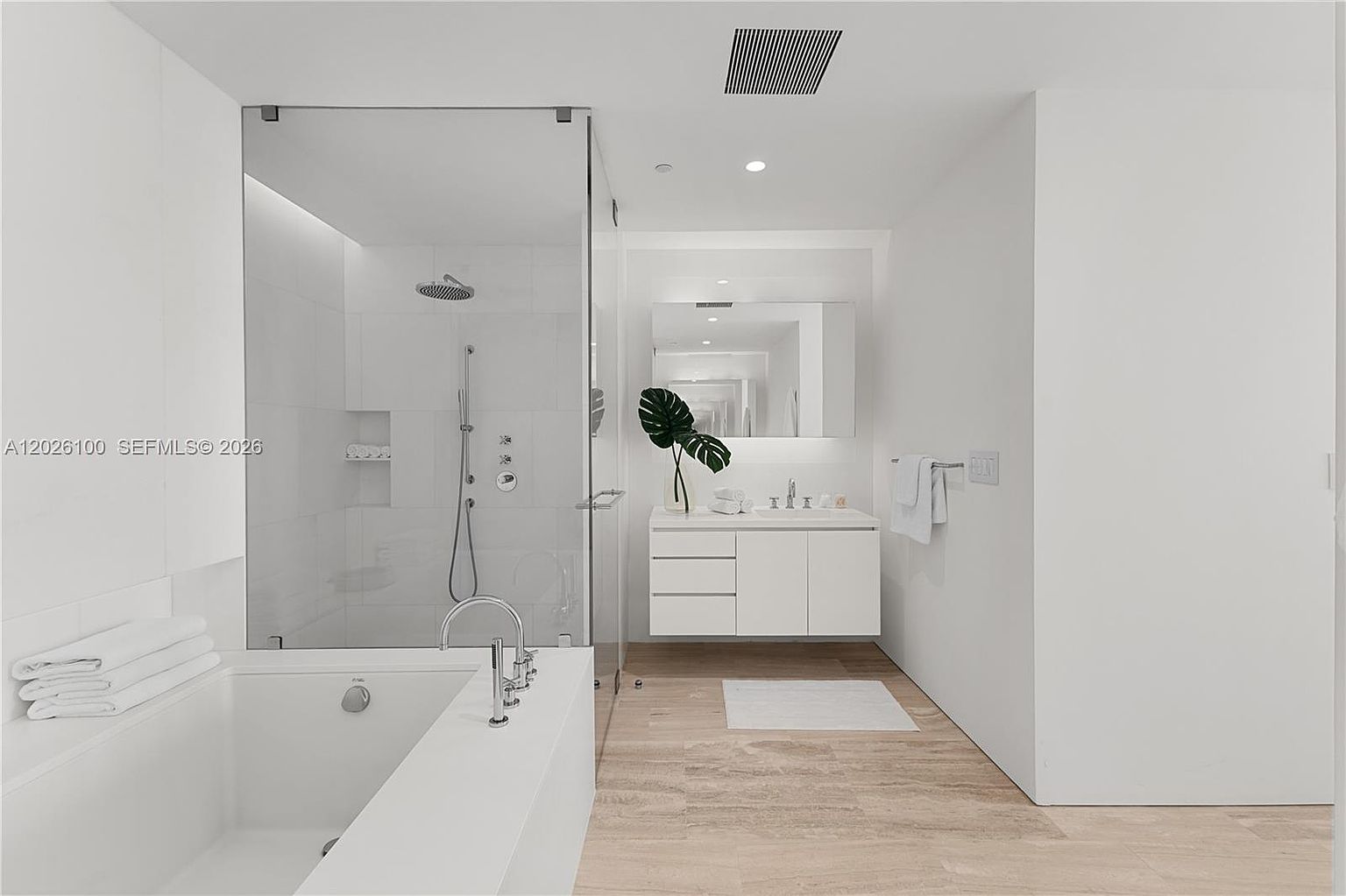 This bright and modern primary bathroom features a clean, minimalist aesthetic with white walls and light-toned wood-look flooring. The space includes a deep soaking tub in the foreground, a glass-enclosed walk-in shower with a rainfall showerhead, and a sleek white vanity with a rectangular sink and a large mirror. The overall impression is one of airy, spa-like tranquility, enhanced by the neutral color palette and contemporary fixtures.