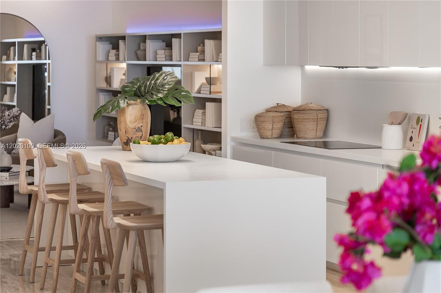 This modern, minimalist kitchen features a sleek white island with light wood bar stools and a decorative bowl of fruit. The space is accented by a tall, open shelving unit filled with books and decor, while a vibrant bouquet of pink flowers adds a pop of color in the foreground. The clean lines and neutral palette create an airy, sophisticated atmosphere perfect for contemporary living.