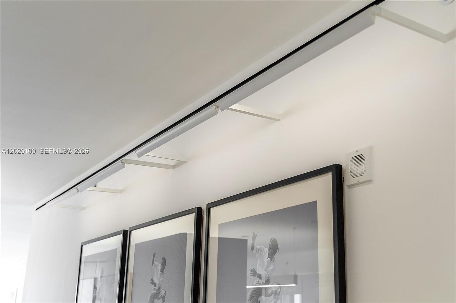 This detail shot highlights a minimalist track lighting system mounted on a clean white wall, positioned above a series of framed black-and-white photographs. A small, square speaker is integrated into the wall surface to the right of the artwork, contributing to a sleek and modern aesthetic. The perspective is angled slightly upward, emphasizing the clean lines of the lighting fixture and the gallery-style presentation of the wall art.