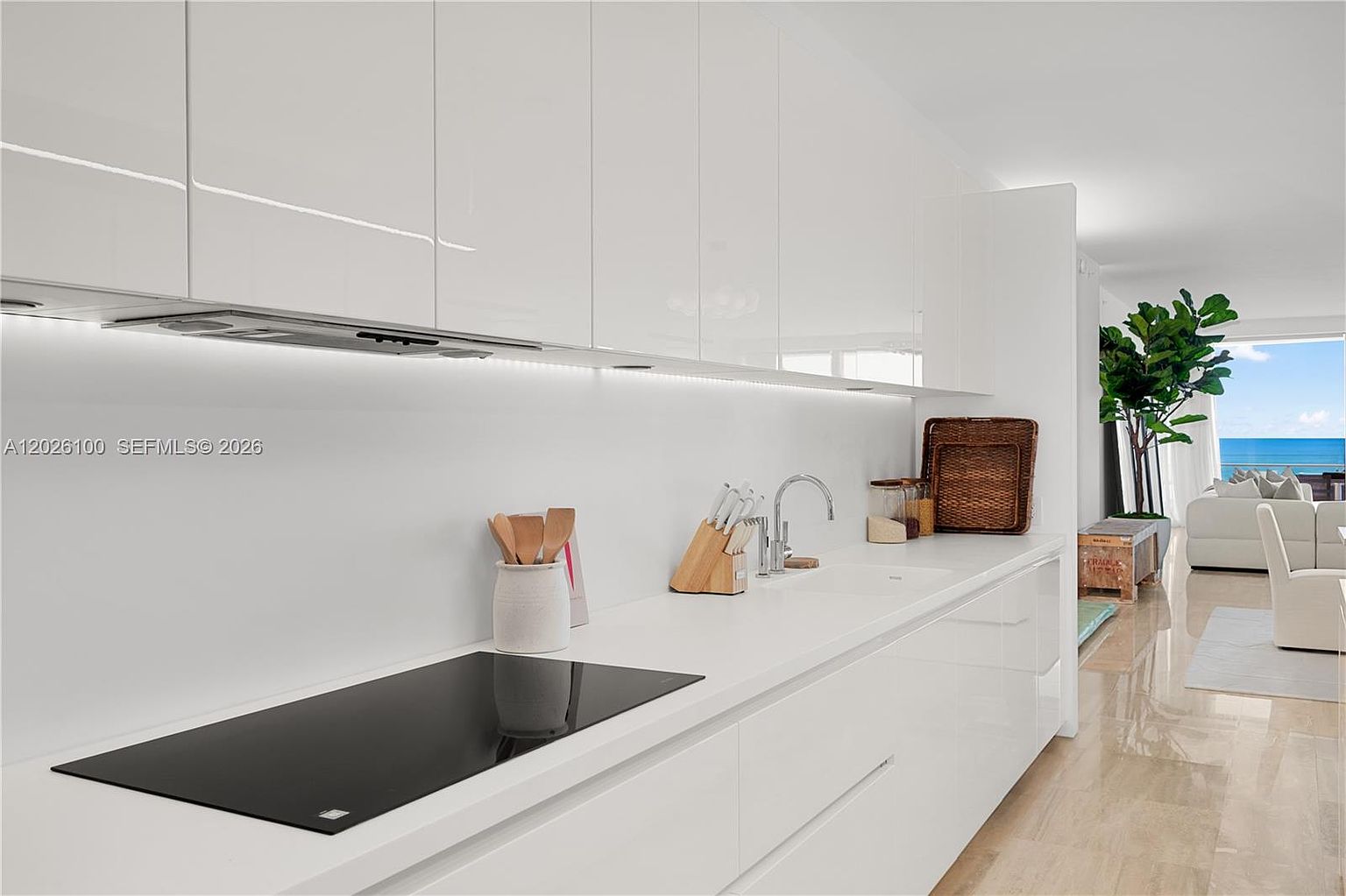 This sleek, modern kitchen features minimalist white cabinetry and a seamless white countertop with an integrated cooktop. The space is illuminated by under-cabinet lighting, creating a bright and airy atmosphere that flows directly into an open-concept living area with a view of the ocean. The perspective is a wide-angle shot from the kitchen counter, emphasizing the clean lines and the transition from the interior workspace to the coastal living space.