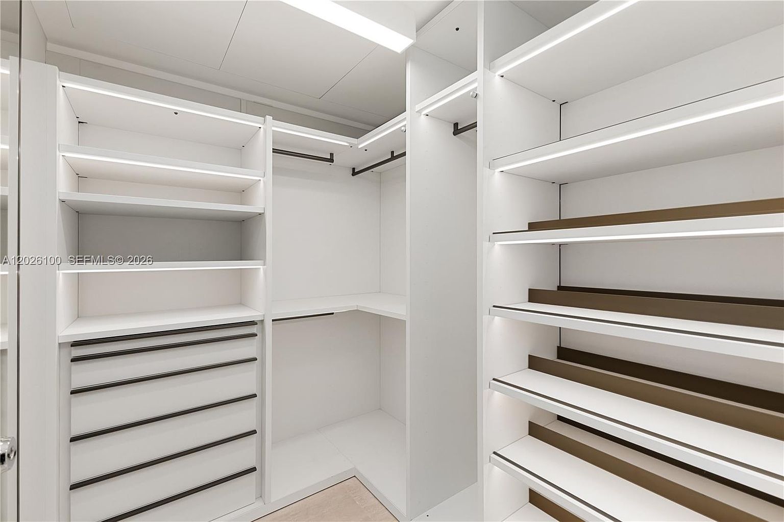 This modern walk-in closet features custom white cabinetry with integrated LED strip lighting along the shelves. The layout includes a combination of open shelving, hanging rods, and a bank of drawers with dark hardware, providing a clean and organized storage solution. The perspective is from the entrance, showcasing the efficient use of space and the bright, minimalist aesthetic.