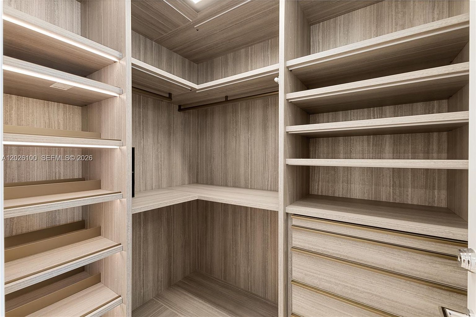 This walk-in closet features custom light wood cabinetry with a clean, modern aesthetic. The space includes a combination of open shelving, hanging rods, and a set of drawers with metallic pulls, providing ample organized storage. The perspective is a straight-on view into the corner of the closet, highlighting the efficient use of space and high-quality built-in design.