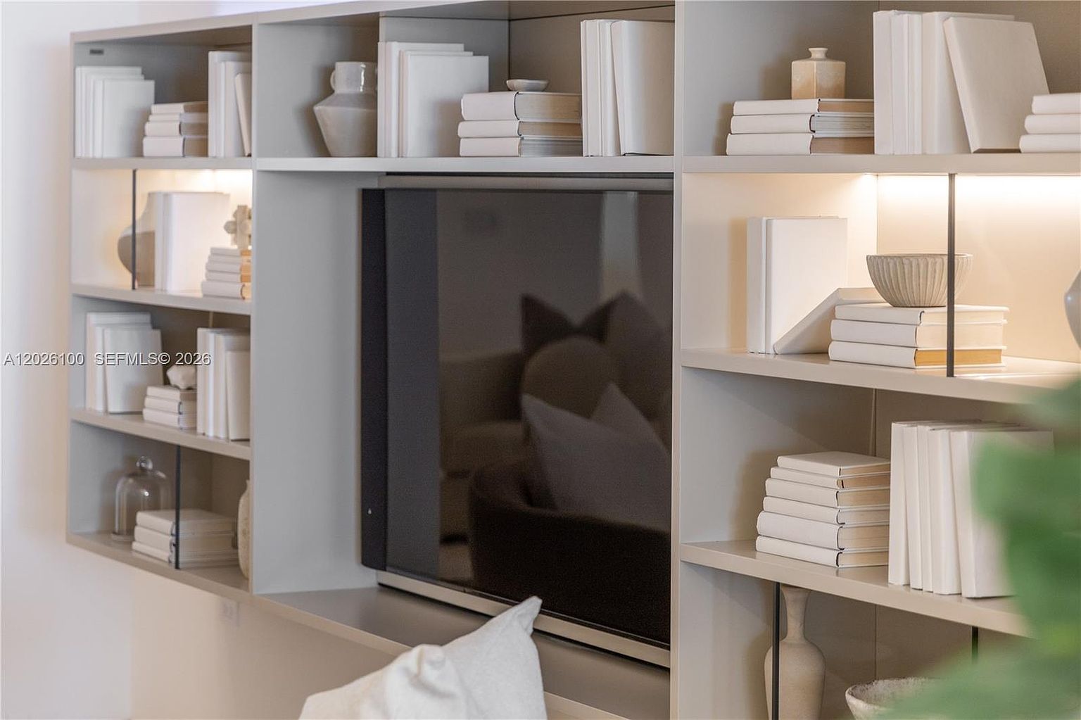 This image showcases a custom-built, light-toned shelving unit integrated into a living room wall, featuring a central recessed space for a television. The shelves are styled with minimalist white books, decorative ceramic vessels, and soft integrated lighting that highlights the curated display. The overall aesthetic is clean, modern, and sophisticated, emphasizing organized storage and a neutral color palette.