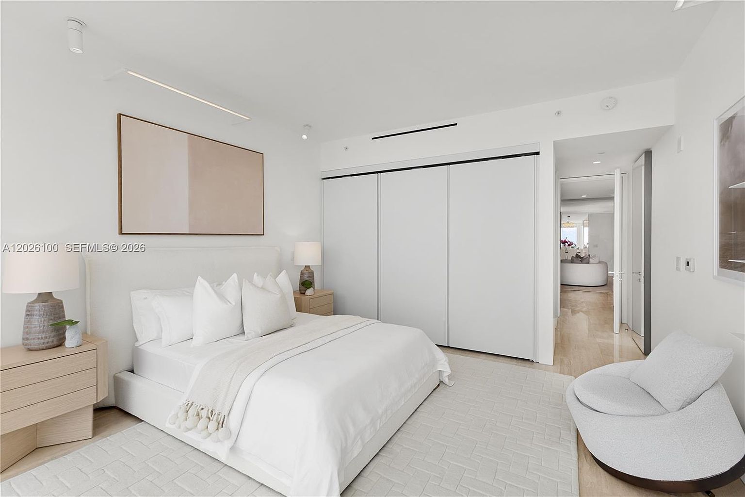 This bright and airy primary bedroom features a minimalist aesthetic with a neutral color palette and clean lines. The space is anchored by a plush bed with white linens, flanked by light wood nightstands and matching lamps, while a modern, curved accent chair sits in the corner. A large, minimalist abstract art piece hangs above the headboard, and a set of white sliding closet doors provides ample storage, leading the eye toward an adjacent room visible through the doorway.