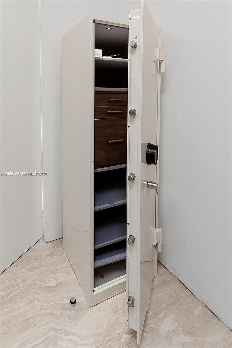 This image features a tall, off-white security safe with its heavy door swung open to reveal a well-organized interior. The inside of the safe is equipped with a set of dark wood drawers and several adjustable shelves, providing secure storage for valuables. The safe is positioned against a plain wall on a light-colored, polished stone floor, highlighting its robust construction and functional design.