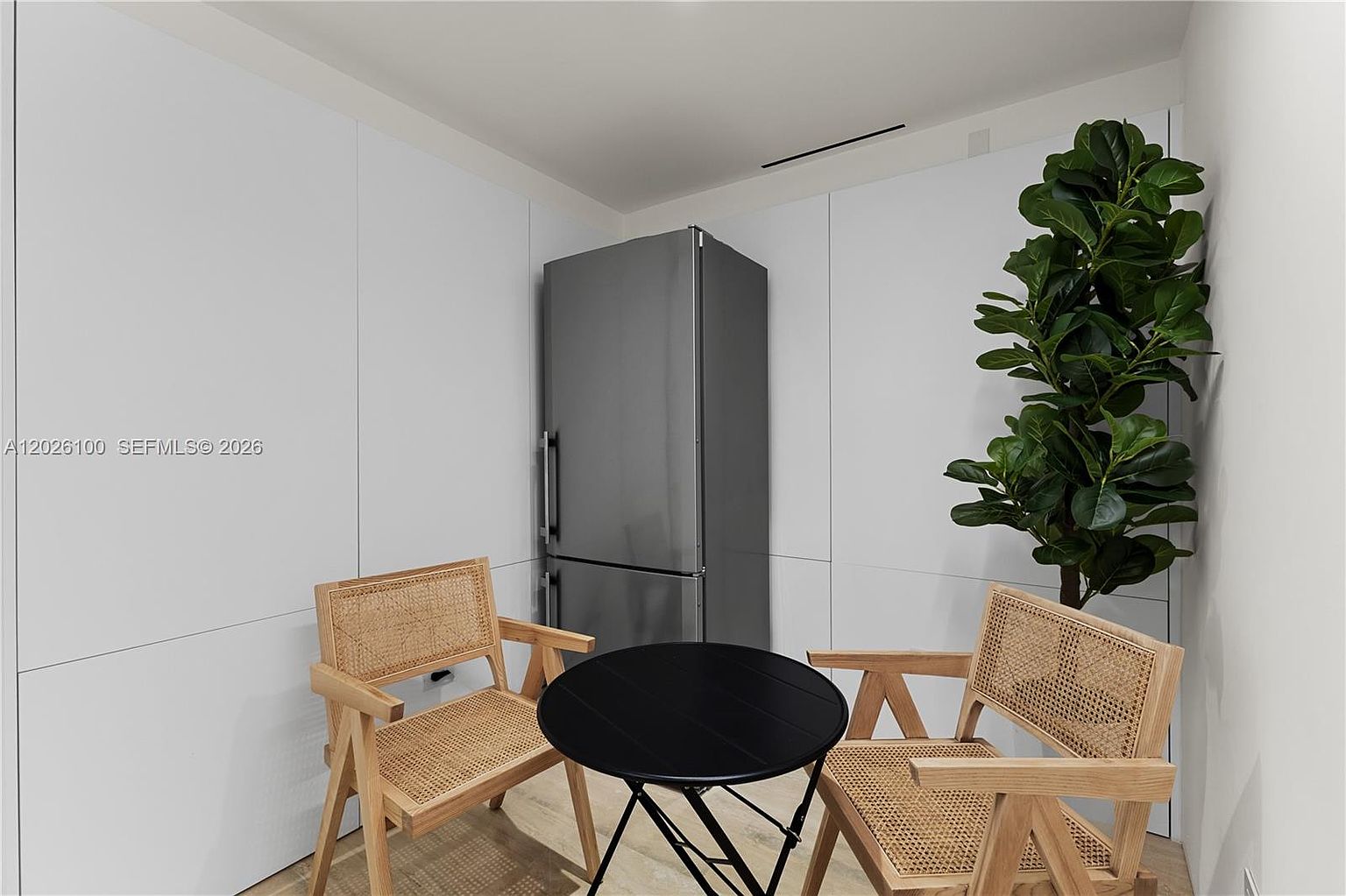 This minimalist kitchen nook features a stainless steel refrigerator integrated into a wall of sleek, white cabinetry. A small, round black table is centered between two light wood chairs with woven cane detailing, creating a cozy breakfast area. A tall, vibrant green potted plant adds a touch of nature to the corner, enhancing the clean and modern aesthetic of the space.