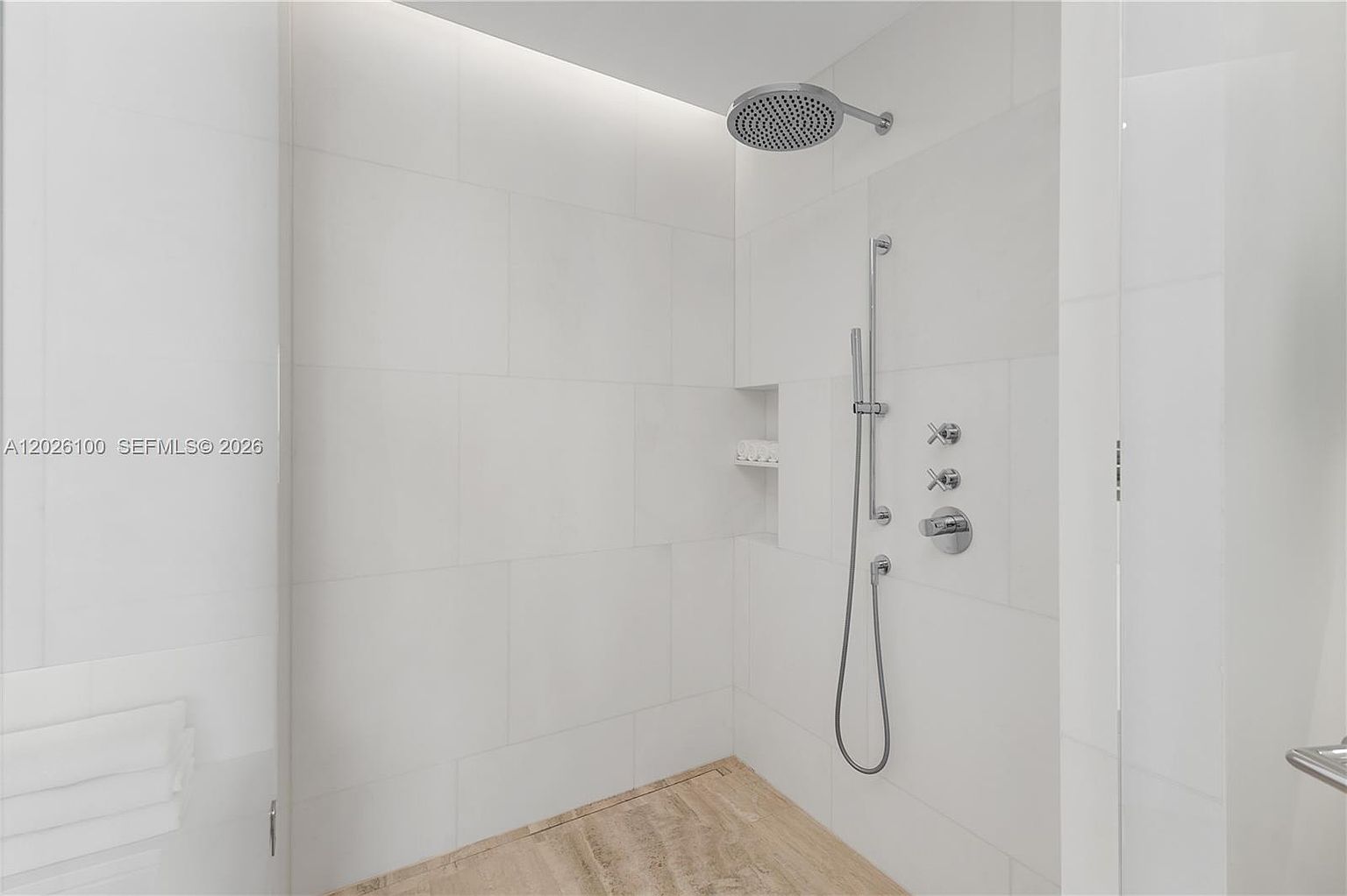 This modern walk-in shower features large-format white wall tiles and a textured, light-toned stone floor. The space is equipped with a circular rainfall showerhead, a handheld sprayer, and a recessed wall niche for storage. The perspective is a straight-on view, highlighting the clean, minimalist aesthetic and high-end fixtures.