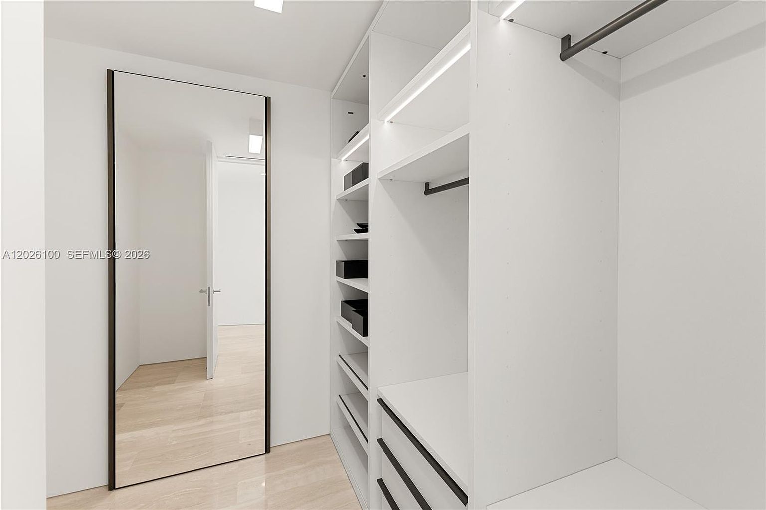 This modern walk-in closet features a minimalist design with crisp white cabinetry, open shelving, and integrated LED lighting. A large, full-length mirror with a dark frame is positioned to reflect the adjacent room, enhancing the sense of space and light. The light-toned flooring and clean lines create a bright, organized, and contemporary aesthetic.