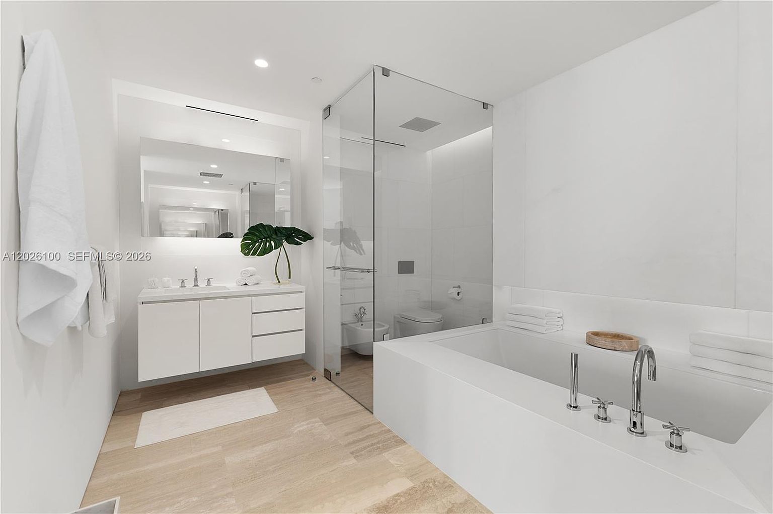 This modern, minimalist bathroom features a sleek white vanity with a single sink, a large wall-mounted mirror, and light wood-toned flooring. A glass-enclosed shower area sits adjacent to a deep soaking tub, creating an open and airy atmosphere. The room is finished with crisp white walls, providing a clean and contemporary aesthetic.