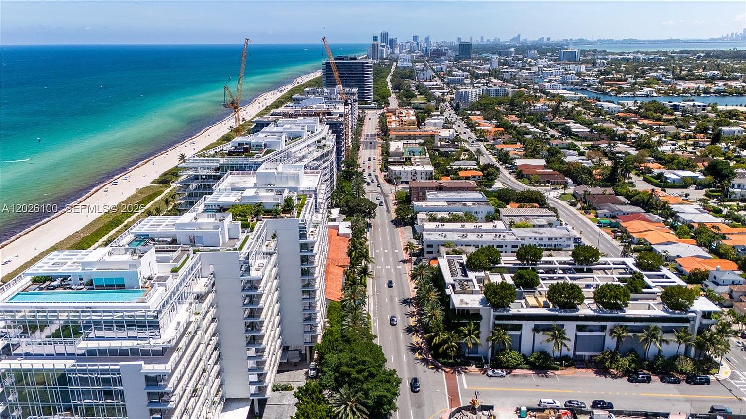 This high-angle aerial view captures a vibrant coastal neighborhood, showcasing a prominent modern residential complex situated directly along a pristine sandy beach and the turquoise ocean. The perspective highlights the urban layout, with a busy thoroughfare running parallel to the coastline, flanked by lush palm trees and various low-rise and high-rise buildings. The scene conveys a sunny, tropical atmosphere typical of a prime beachfront real estate location.
