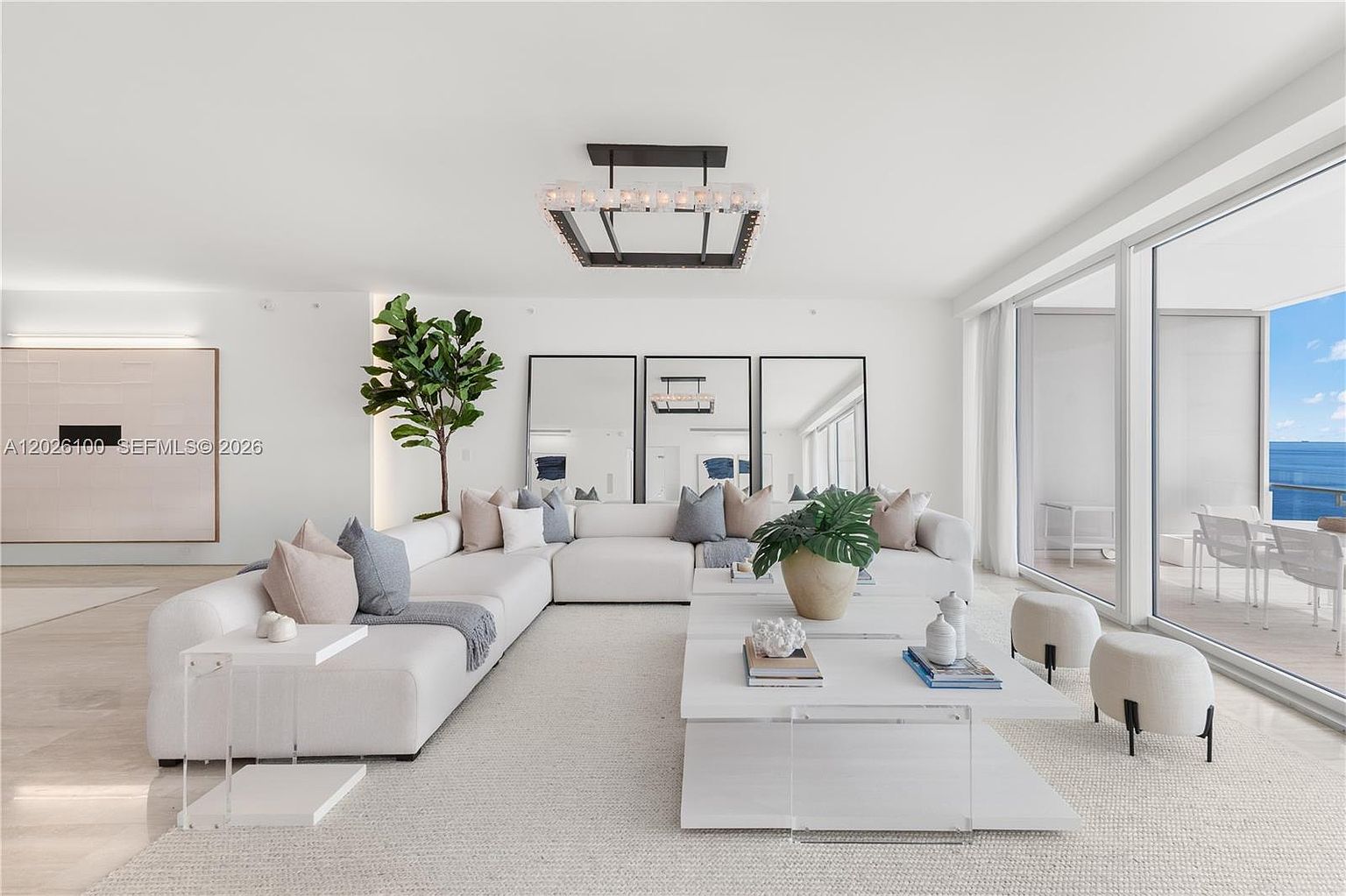 This bright and airy living room features a large, neutral-toned sectional sofa arranged around a tiered white coffee table. The space is defined by its minimalist aesthetic, highlighted by floor-to-ceiling windows that offer expansive ocean views and a modern geometric chandelier. Three large mirrors behind the sofa enhance the sense of depth and light, creating a sophisticated and serene atmosphere.