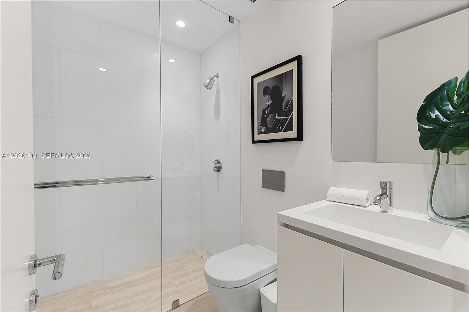 This modern, minimalist bathroom features a sleek wall-mounted toilet and a glass-enclosed walk-in shower with light-toned tile flooring. A white vanity with a rectangular sink sits beneath a large, frameless mirror, complemented by a decorative plant and a framed piece of art on the wall. The space is bright and clean, utilizing a neutral color palette to create a contemporary and airy atmosphere.