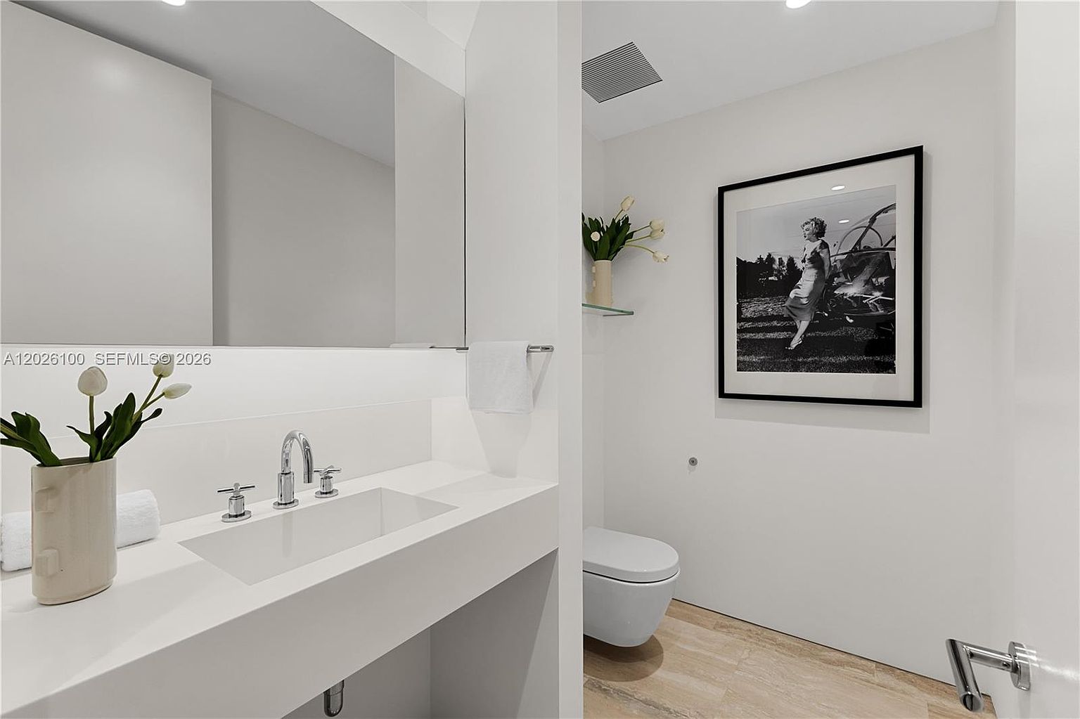 This modern powder room features a minimalist aesthetic with a sleek, wall-mounted vanity and a rectangular vessel sink. The space is accented by a black-and-white framed photograph on the wall and a small vase of white flowers, creating a clean and sophisticated atmosphere. The light-toned flooring and neutral walls enhance the bright, airy feel of this contemporary bathroom.