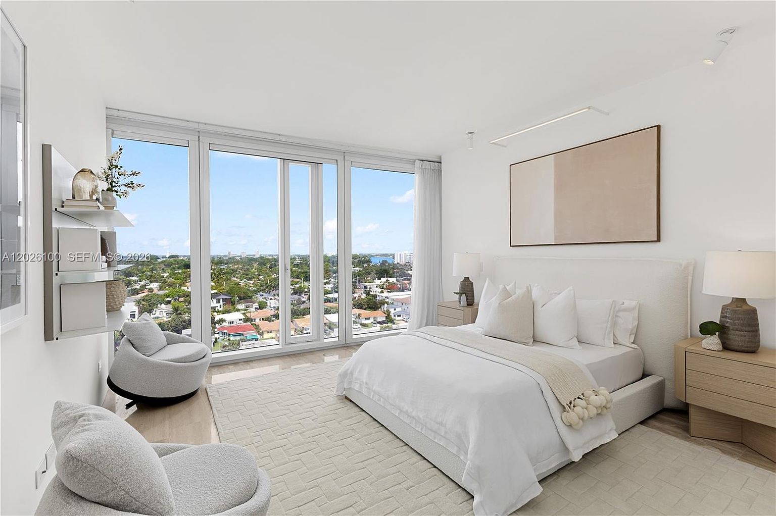 This bright and airy primary bedroom features a neutral color palette with a plush white bed and matching textured area rug. Large floor-to-ceiling windows provide an expansive view of the surrounding neighborhood and sky, while two modern armchairs offer a comfortable seating area. The room is accented by minimalist wall art and warm bedside lighting, creating a serene and sophisticated atmosphere.
