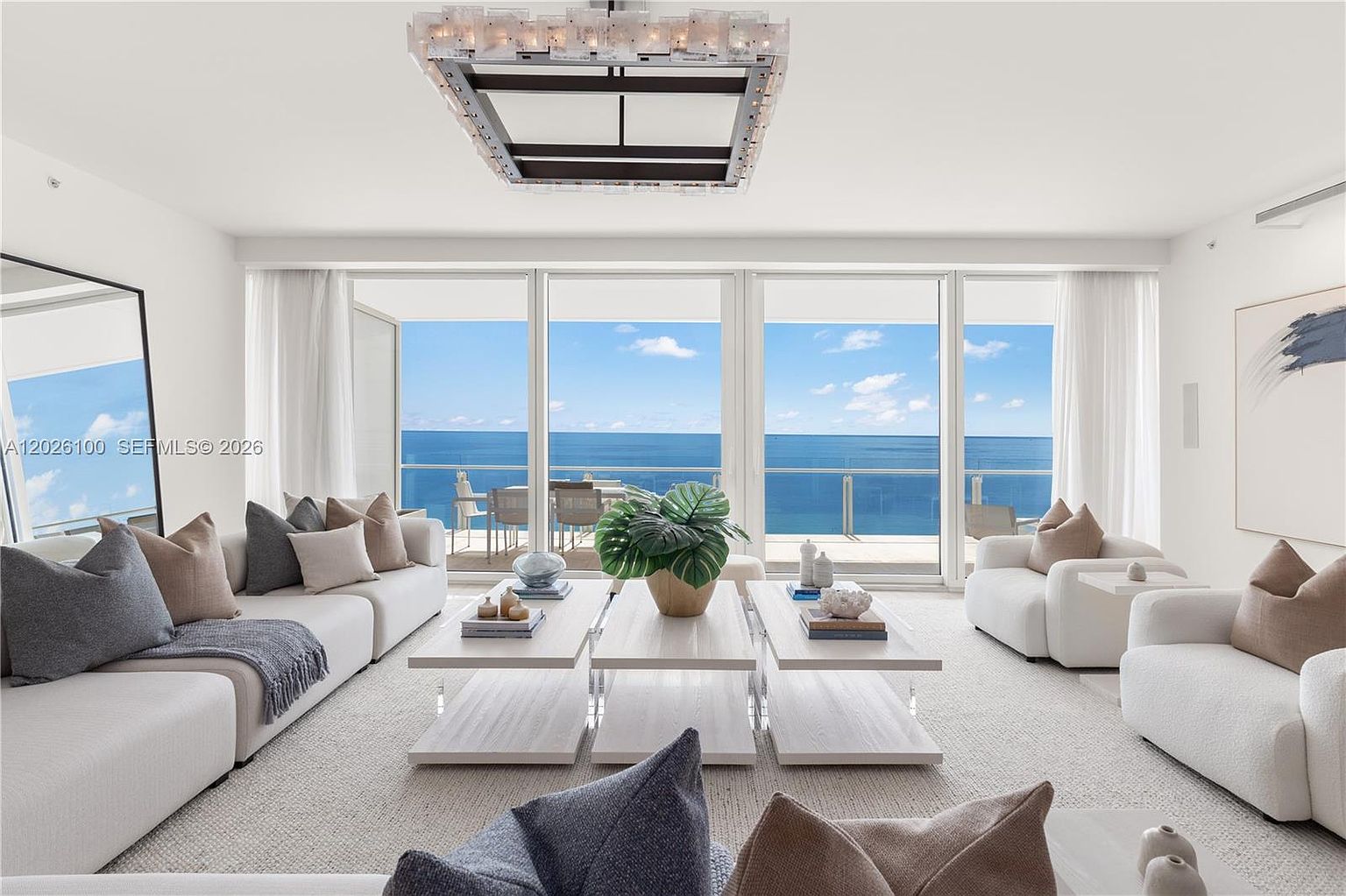 This bright and airy living room features a neutral color palette with plush white sofas and a pair of matching armchairs arranged around a set of low-profile, light-toned coffee tables. The space is anchored by a large, modern chandelier and offers expansive, unobstructed views of the ocean through floor-to-ceiling glass walls that lead to a balcony. The overall aesthetic is sophisticated and serene, emphasizing a seamless connection between the luxurious interior and the coastal horizon.