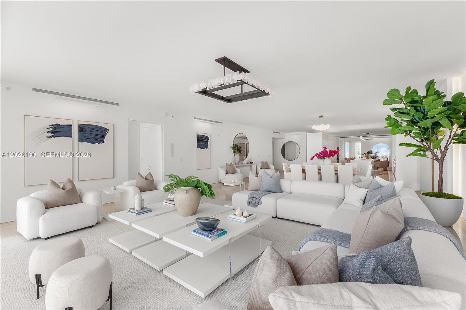 This expansive, open-concept living area features a bright, minimalist aesthetic with a large white sectional sofa and matching armchairs arranged around a cluster of low-profile square coffee tables. The space is accented by a large potted fiddle leaf fig, abstract wall art, and a contemporary geometric chandelier, all set against a neutral color palette. The wide-angle perspective captures the seamless flow from the seating area toward the dining space and beyond, emphasizing the airy and sophisticated atmosphere of the home.