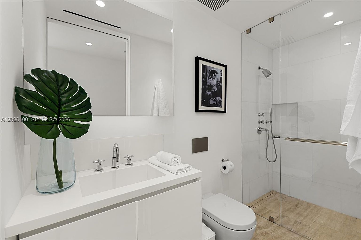 This modern, minimalist bathroom features a sleek white vanity with a rectangular sink and polished chrome fixtures. A glass-enclosed walk-in shower with light-toned tile flooring and wall tiling creates an open, airy feel. The space is accented by a large mirror, a framed black-and-white photograph, and a decorative plant, contributing to a clean and sophisticated aesthetic.