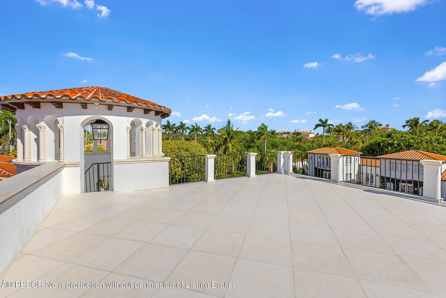 This expansive rooftop terrace features light-colored stone tiling and a classic Mediterranean-style rotunda with arched openings and a terracotta-tiled roof. A black metal railing surrounds the perimeter, offering an elevated vantage point overlooking lush tropical landscaping and neighboring villas. The bright, sunny atmosphere and open layout create an ideal space for outdoor entertaining or relaxation.