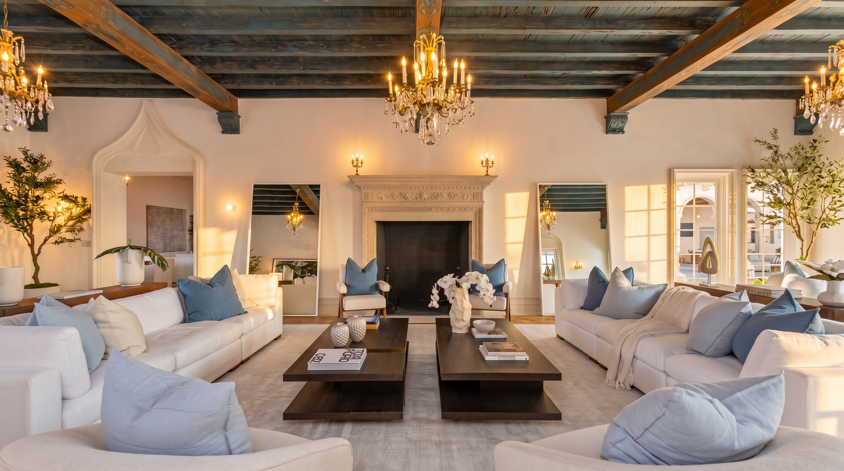 This elegant living room features a symmetrical layout centered around a grand stone fireplace with an ornate mantel. The space is defined by a striking blue-painted wood-beamed ceiling, plush white sofas with blue accent pillows, and two large rectangular coffee tables. Two tall mirrors flank the fireplace, reflecting the room's sophisticated decor and crystal chandelier, creating a bright and airy atmosphere.