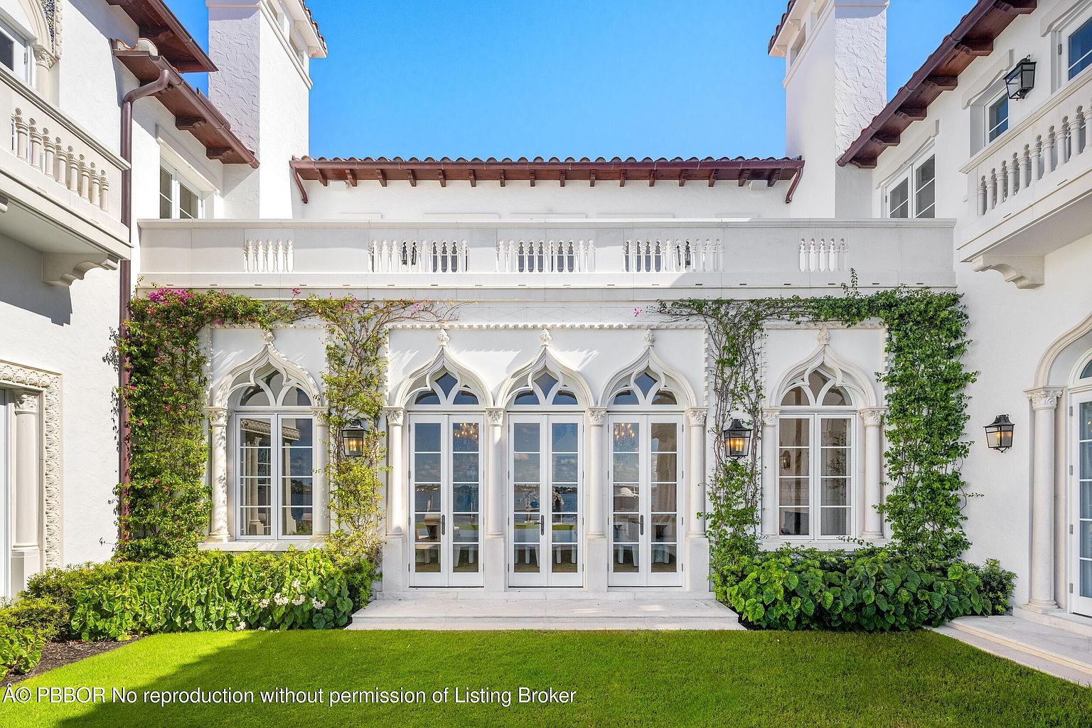 This elegant rear exterior view showcases a Mediterranean-inspired estate with intricate Gothic-style arched windows and glass doors. The white stucco facade is adorned with lush climbing vines, and a stone balustrade terrace sits above the ground-level patio. A manicured green lawn stretches across the foreground, creating a serene and sophisticated outdoor living space.